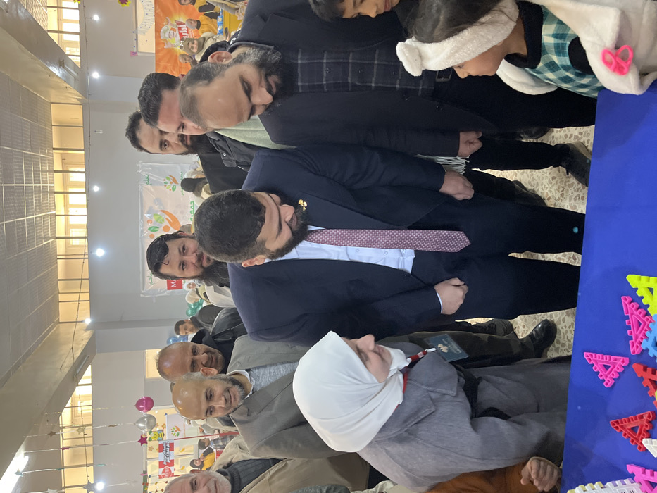 An indoor scene in Kafr Batna, Syria, depicts a group of individuals gathered around a blue table displaying colorful plastic interlocking building toys. In the immediate foreground, a man in a dark blue suit with a light blue shirt and a patterned red and white tie, adorned with a gold lapel pin, is bending forward, looking down at the toys. To his left, a woman wearing a light gray coat and a white hijab smiles while also leaning forward, observing the table. She wears a red and white lanyard with an identification badge. To the right of the man in the suit, another man with a dark beard and a black jacket over a checkered shirt leans forward, directing his gaze downwards. A small girl, partially visible on the far right, wears a white fluffy headband and a green plaid dress, looking up towards the suit-wearing man. Behind this foreground group, several adult men are visible, some with beards, dressed in various casual and semi-formal attire, observing the activity. One man with a bald head and beard, wearing a light brown jacket, is smiling. The background features light-colored tiled flooring, fluorescent lighting, and decorated walls with star garlands, pink and silver balloons, and banners with Arabic text and logos, one prominently displaying a hand reaching towards a green leaf, indicative of an organized event or community gathering.