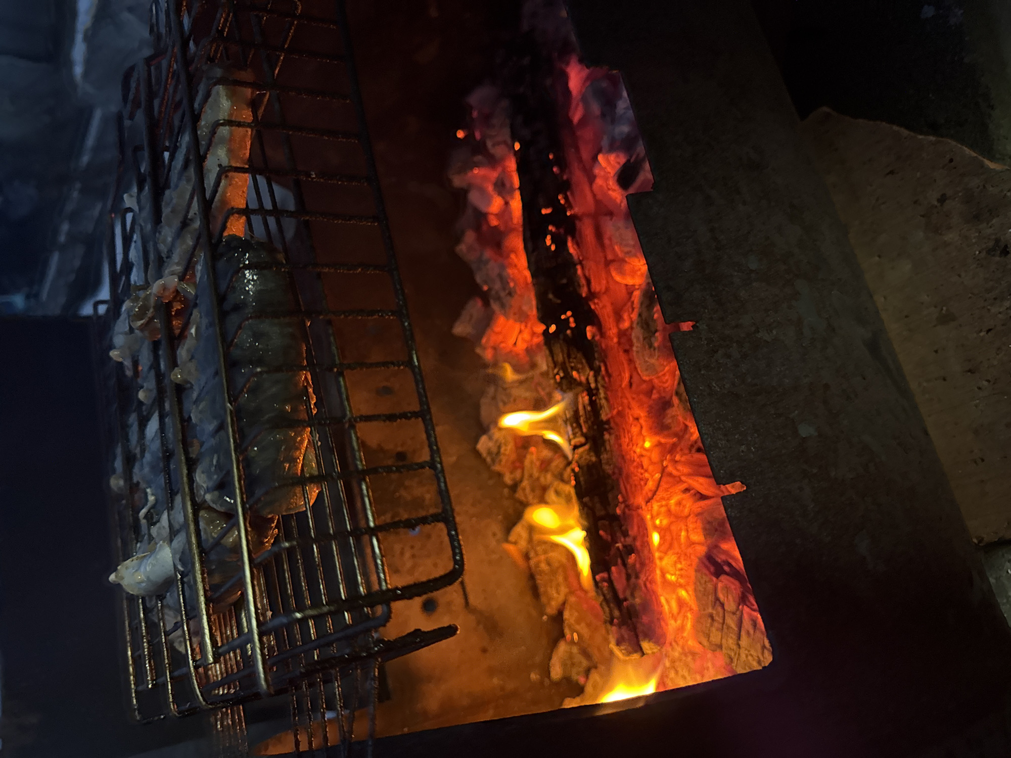 The image shows food being grilled over a wood fire. The food is contained within a wire grilling basket. Flames and burning wood are visible next to the basket. The grill apparatus appears to be made of metal. The photograph was taken in Chaikovsky, Russia.