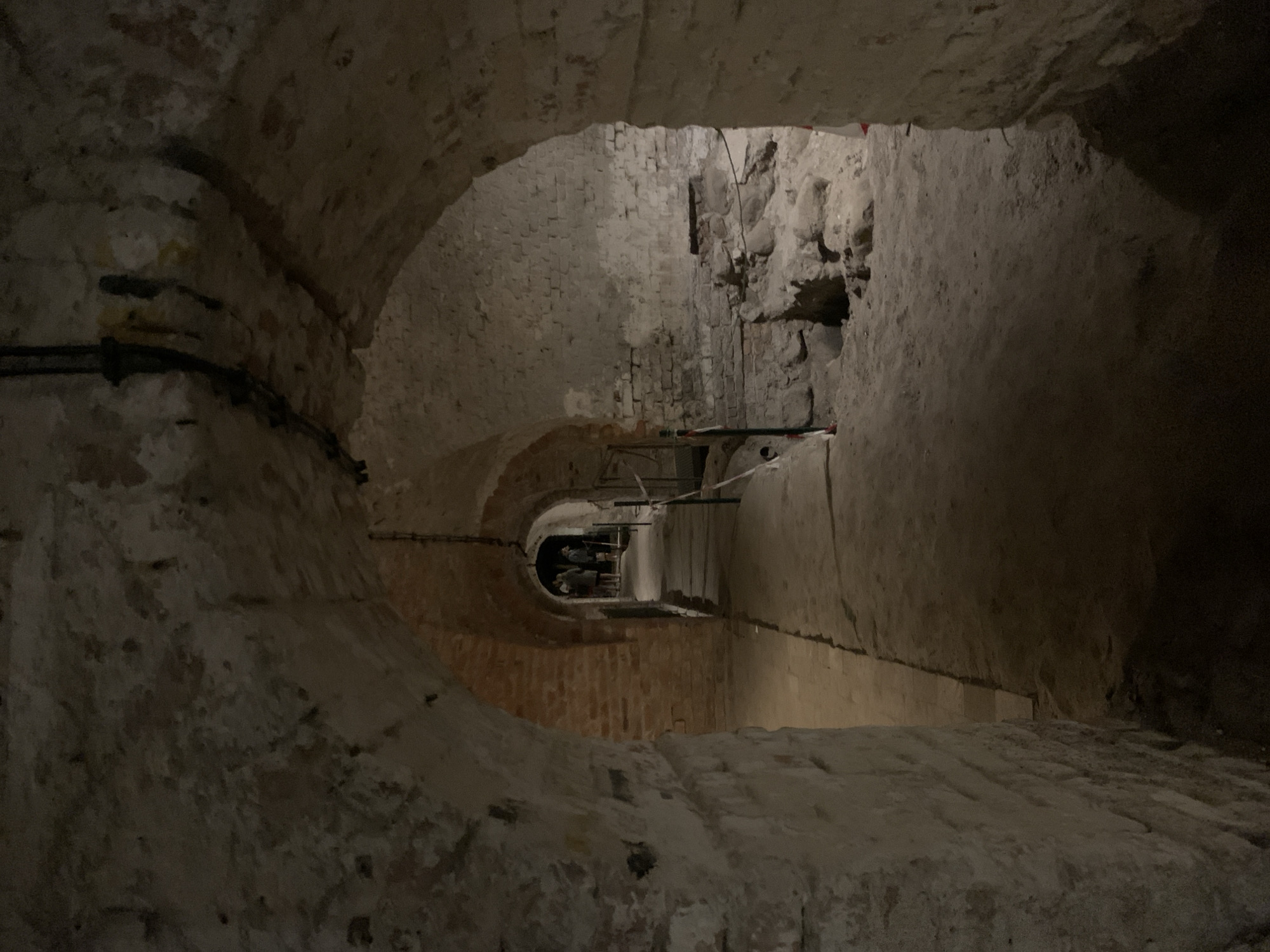 The image depicts a dimly lit, arched brick corridor within an underground space. The walls are constructed of rough-hewn bricks and some stone, showing signs of age and repair. The floor is covered in a layer of dirt and sand. Several arches lead deeper into the corridor, creating a sense of depth. In the distance, a group of people are visible, appearing to be tourists or visitors, walking down the corridor. Red and white tape, likely used to cordon off areas or guide visitors, is visible along the path. The overall impression is of an archaeological site or an old underground structure, potentially a cellar or catacombs. This scene is located in Poznan, Poland, specifically within the subterranean route of the Poznan Old Market Square.