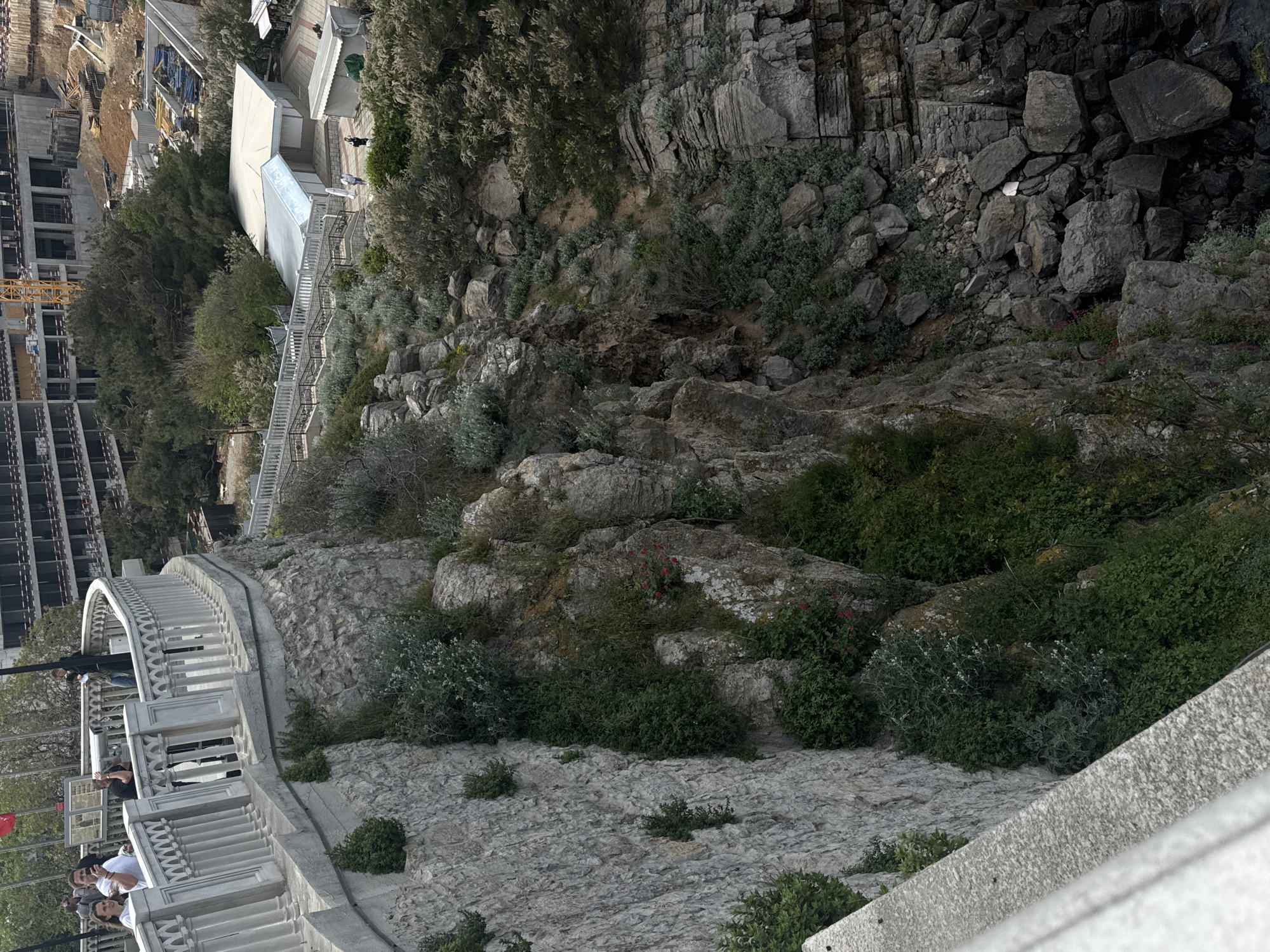 A high-angle view, rotated counter-clockwise, features a stone wall with a white balustrade along its top edge, occupied by several people. The wall descends sharply into a rocky terrain covered with vegetation and a steep cliff face on the right. A construction site with a crane and unfinished buildings is visible on the left side of the image, along with a white building with a staircase. The scene suggests an urban area adjacent to natural cliffs, likely the city of Гаспринский поселковый совет, Ukraine. The people are standing on the structure, seemingly observing the view.