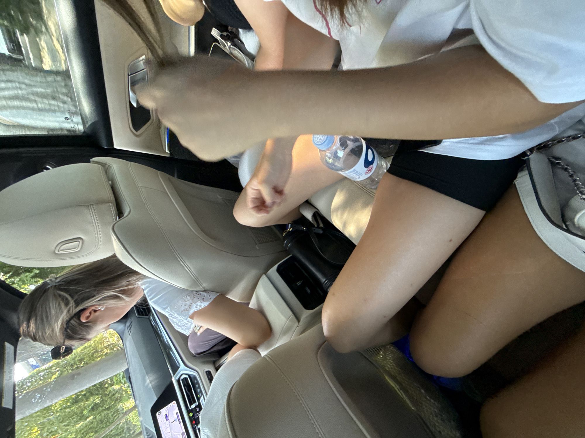 The image captures the interior of a modern car, likely a BMW, seen from the perspective of someone in the back seat. The upholstery is a light beige leather. In the foreground, the legs of two individuals are visible. One person, wearing black shorts and a white top, has their legs crossed, with a bottle of water and a handbag beside them. Another person's legs, in black shorts and a white top, are also visible. In the middle ground, a woman with blonde hair pulled back, wearing a white top, is seated in the front passenger seat and looking down at her phone. She is wearing sunglasses on top of her head and pearl earrings. The car's dashboard and infotainment screen are visible, displaying a navigation interface. The exterior environment, seen through the windows, suggests a tree-lined road, indicating a journey outside of dense urban areas. There are no specific markers in the image that would definitively place the scene in Chișinău, Moldova.