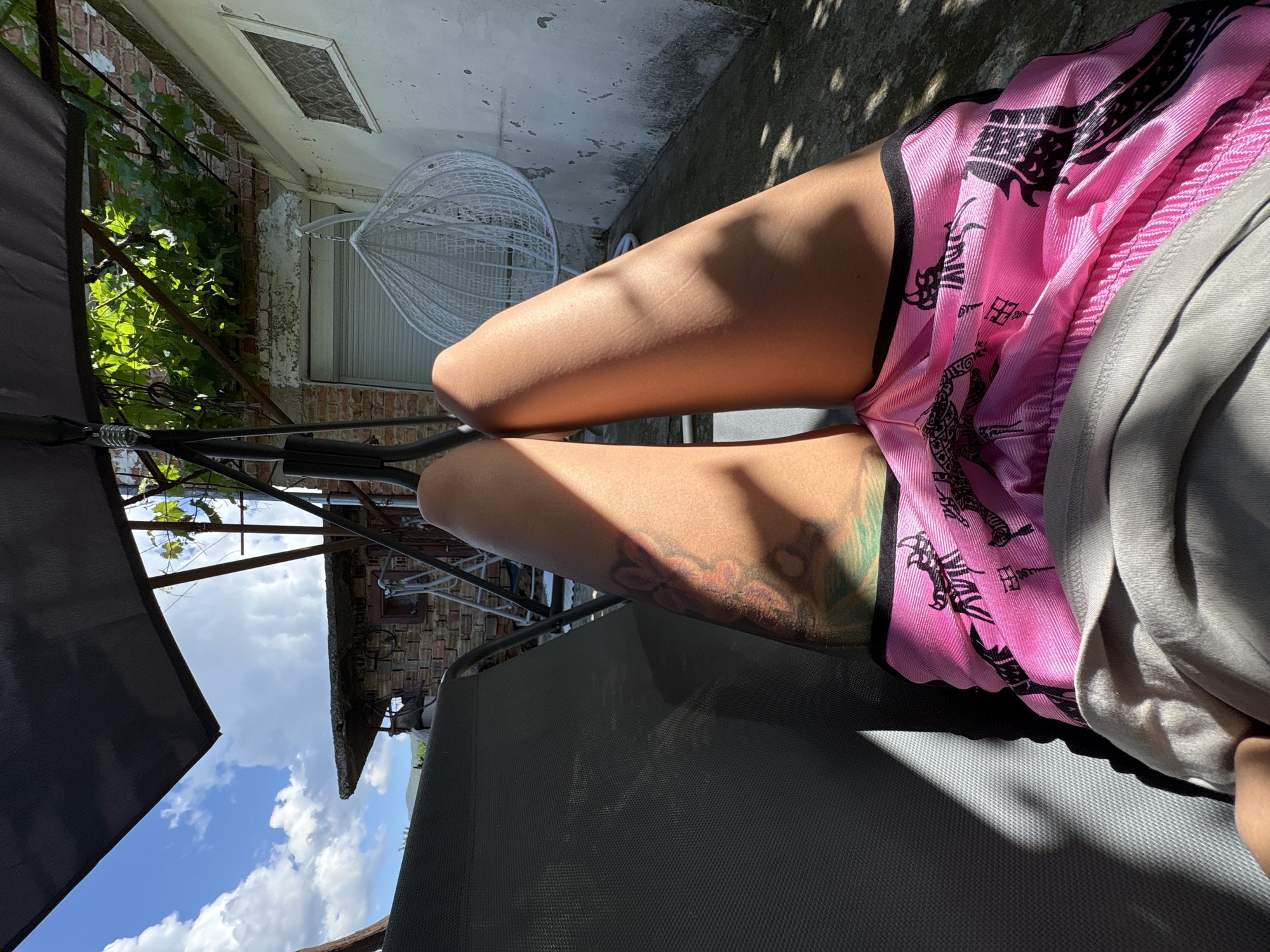 The image shows a person lying on a lounge chair on a sunny day in the backyard of a house. The person is wearing pink shorts with a black pattern and a grey t-shirt. There is a tattoo visible on their leg. The backyard has a brick wall, a white-washed wall with a small window, and a white hanging chair. A swing set with a dark fabric canopy is also present. Green vines are growing on a trellis above the brick wall. The ground is paved with concrete and has shadows cast from the surrounding objects. The image captures a moment of relaxation in a residential outdoor setting.