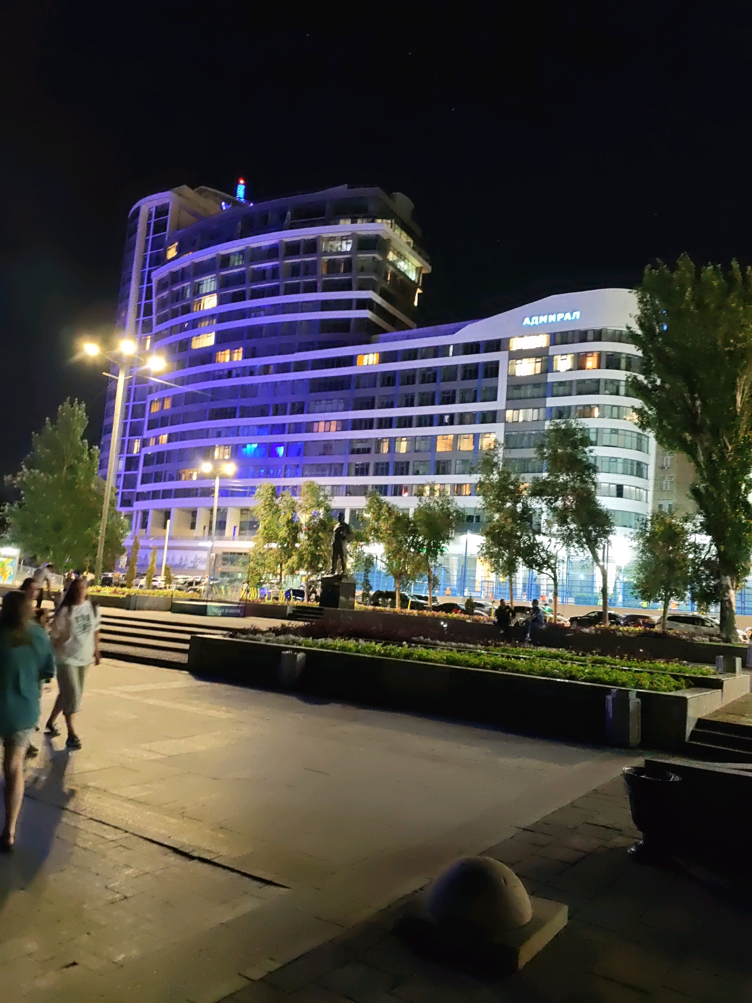 The image depicts the "Admiral" building, a prominent landmark in Rostov-on-Don, Russia, at night. The building's facade is illuminated with vibrant blue lights, contrasting with the warm glow of interior lights visible through its numerous windows. The architectural style appears modern, with curved lines and multiple levels. In the foreground, several individuals are walking on a paved area, their figures silhouetted against the streetlights. Some are in casual attire, suggesting leisure activity. Trees are planted along a landscaped area in front of the building, adding a touch of nature to the urban setting. The scene conveys a sense of a lively city at night, with a blend of modern architecture, pedestrian activity, and urban landscaping.