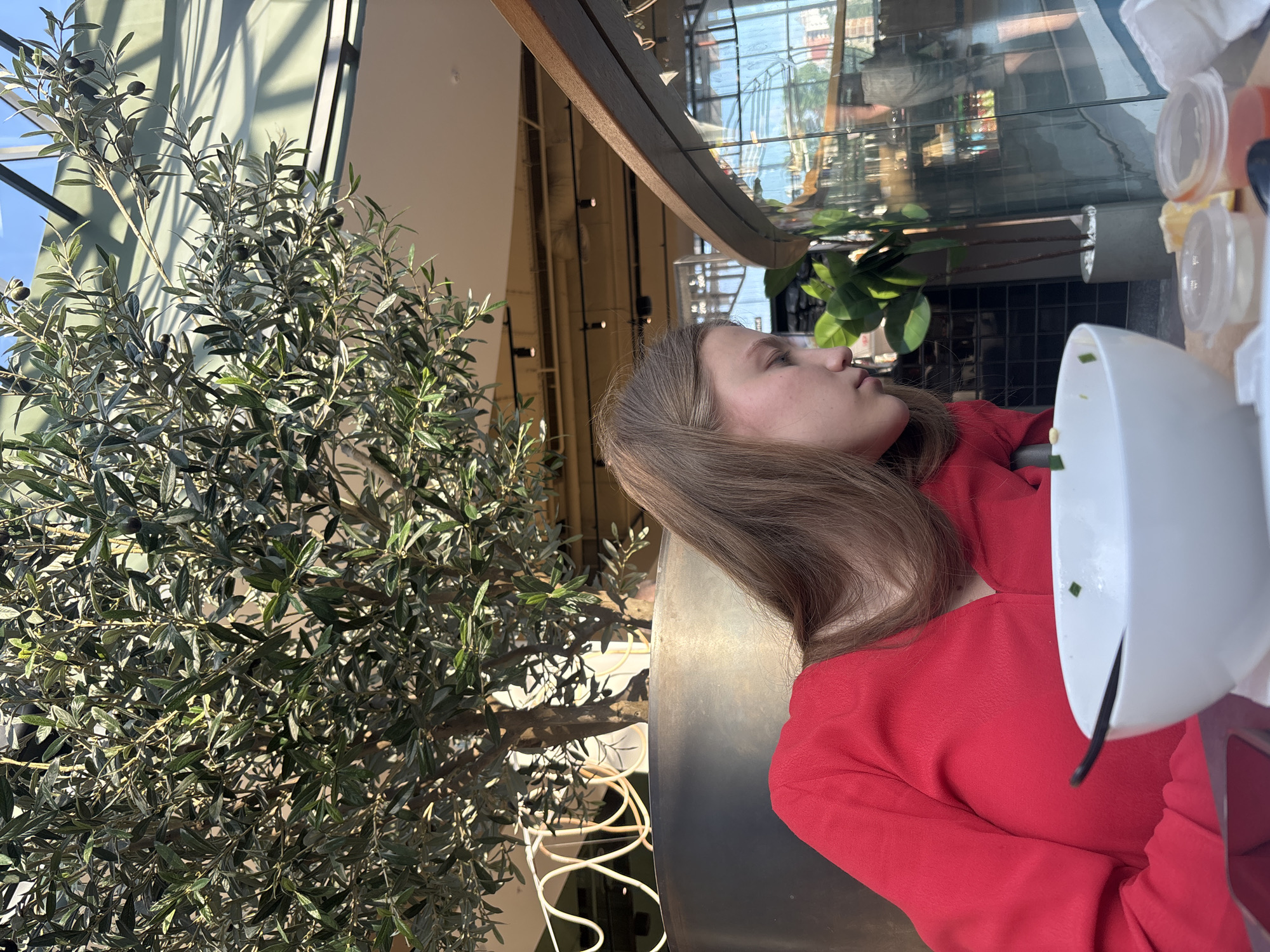 A medium-shot image shows a young woman sitting in a restaurant. She has long brown hair and wears a red shirt. She is looking slightly upward and toward the left. In front of her is a white bowl, and several small containers with condiments are visible in the background. To the left, a large olive tree stands in a planter. The restaurant appears to be in a modern building with a glass ceiling and is located in Perm, Russia.