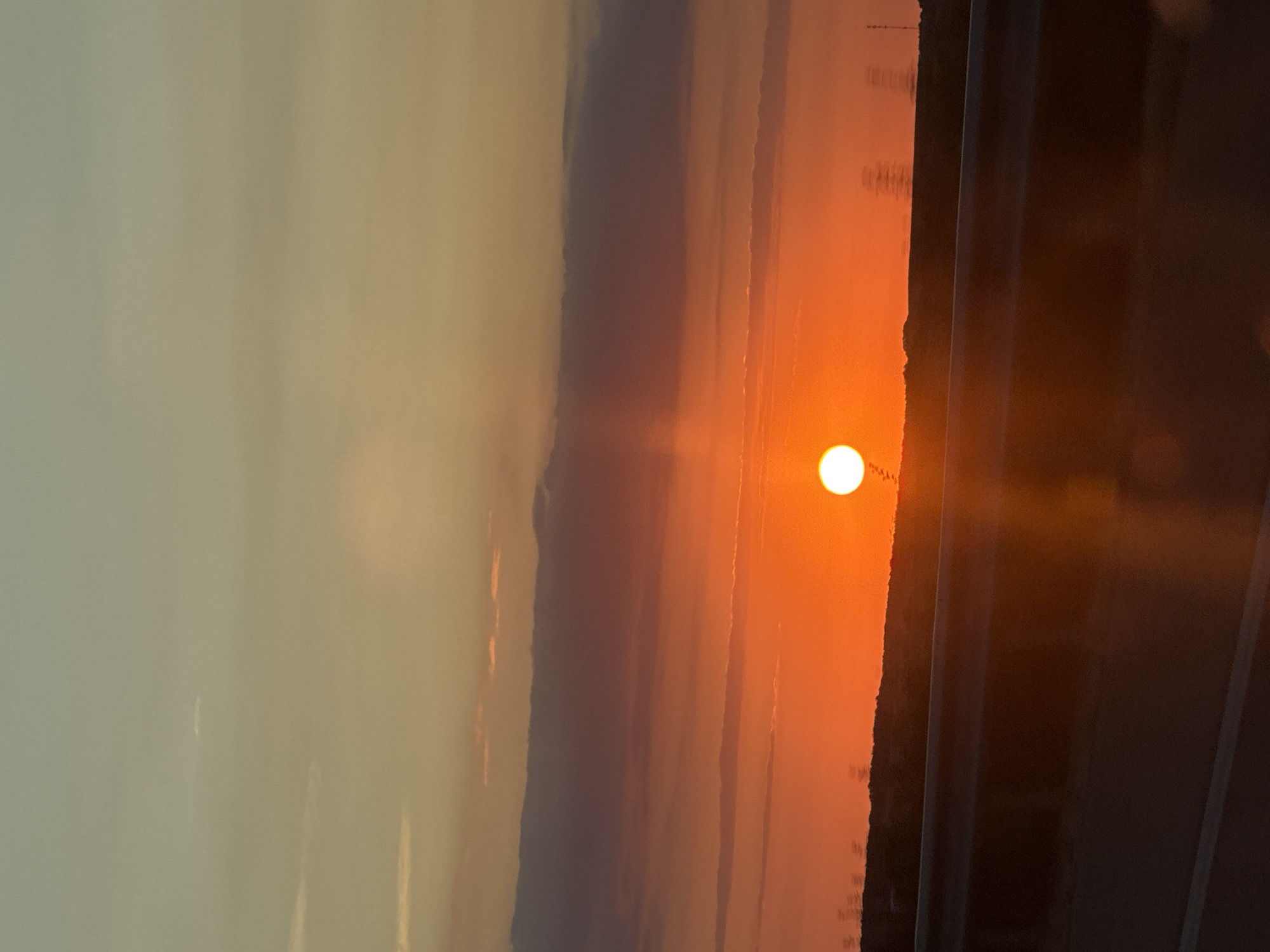 The image captures a sunset viewed from a highway, likely in or near Malko Tarnovo, Bulgaria. The sky is a vibrant gradient of orange and yellow, with scattered clouds. The sun, a bright orb, is setting near the horizon, casting a warm glow over the landscape. A silhouetted line of trees and some structures are visible against the fiery sky. In the foreground, a dark barrier, possibly a highway guardrail, and a section of asphalt road are present. The overall scene is serene and evocative of a journey through the Bulgarian countryside at dusk.