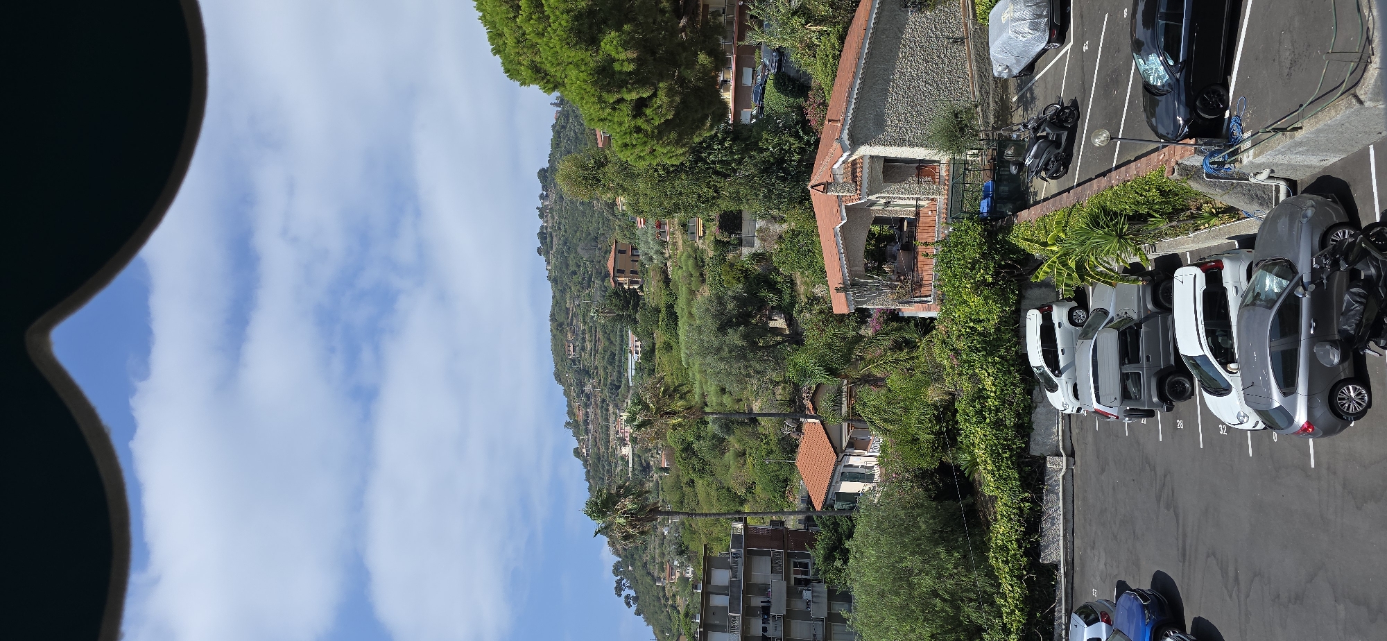 The image shows a view of a hillside residential area with a parking lot in the foreground. A multi-story apartment building is on the left, with several cars parked on asphalt in front of it. Beyond the parking lot, several houses are nestled into the lush green hillside, which is covered in trees and vegetation. Some of the houses have red tile roofs. The sky is blue with scattered white clouds. In the distance, the hillside rises to a peak dotted with more houses. The scene appears to be a sunny day in Sanremo, Italy.