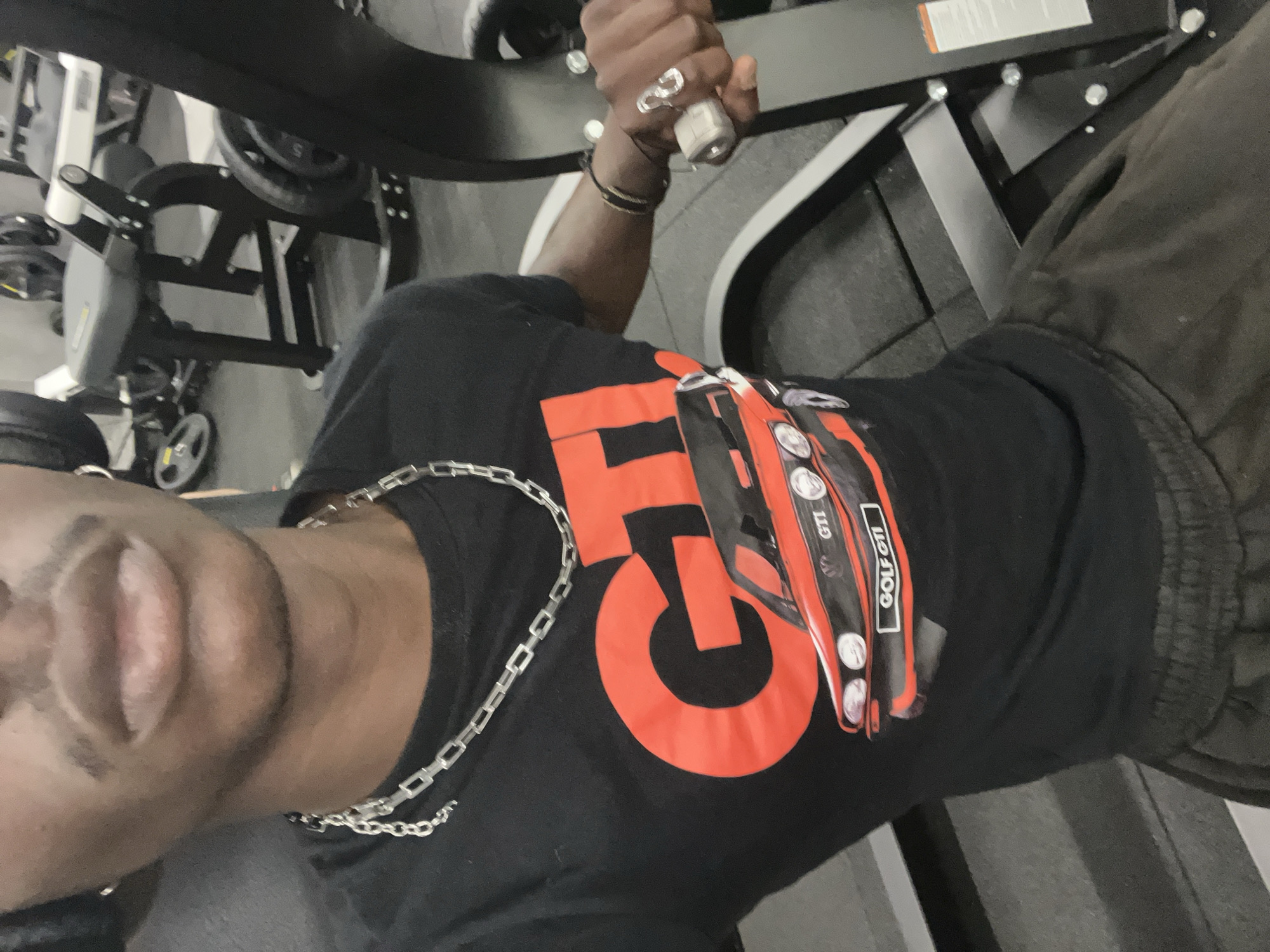 A man is using a weight machine in a gym. He is wearing a black t-shirt with a red "GTI" graphic, a silver chain necklace, and rings on his fingers. He is holding the handle of the weight machine. The gym equipment is black and silver. The background is blurry, showing other gym equipment. The photo is taken at a close angle. There is no information to suggest the location is in Barcelona, Spain.
