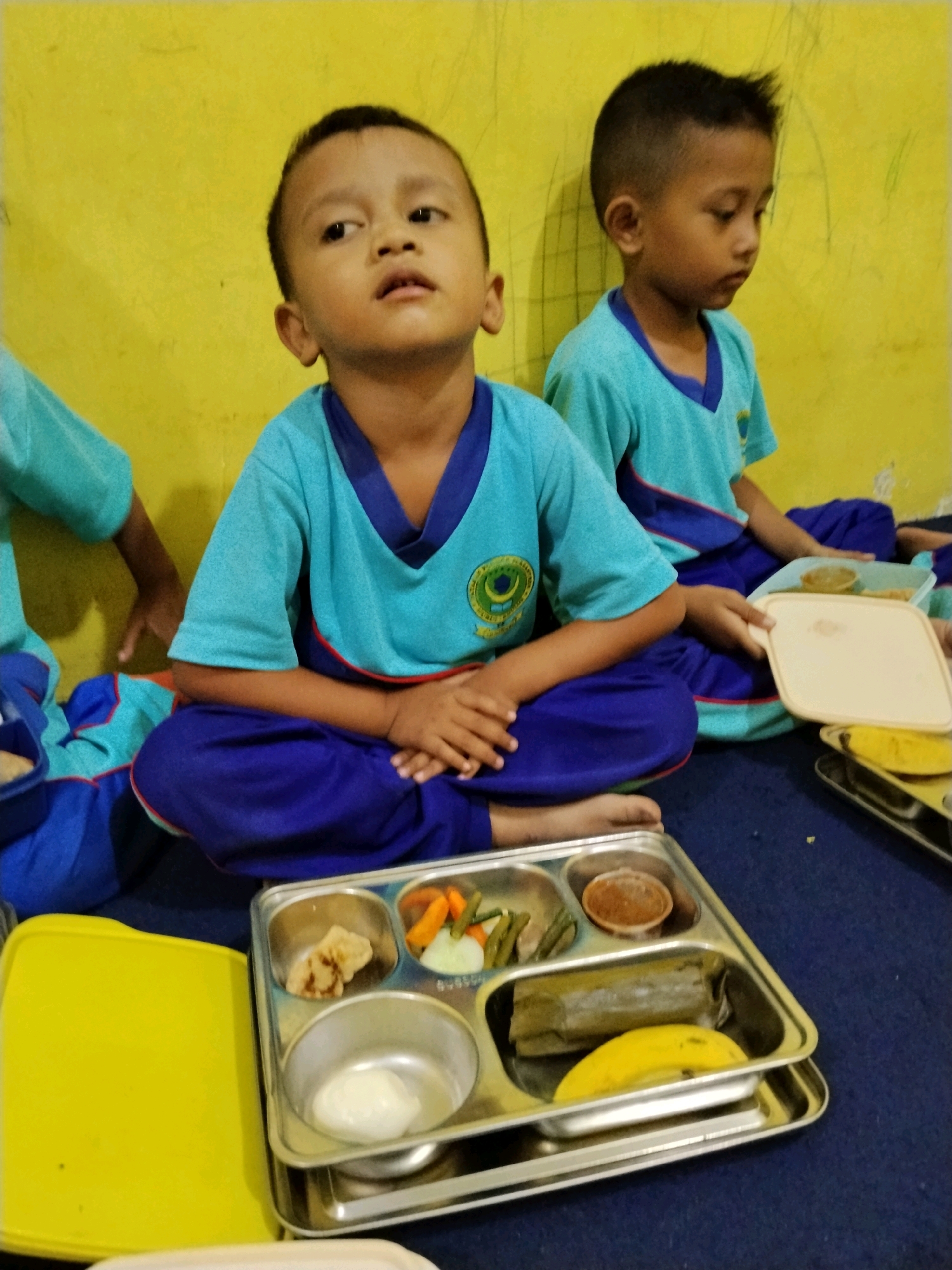 Two young boys, likely students at a school in Panggungan, Indonesia, are seated on the floor in front of a yellow wall, wearing similar blue and teal school uniforms. The boy in the foreground is looking upwards, while the boy behind him is looking down. In front of them are metal lunch trays containing food. The foreground tray holds a piece of fried chicken, some vegetables, a small container of sauce, a wrapped rice dish, and a banana. A yellow lid is to the left of the tray, and another white lid is in the boy's hands on the right. The floor is covered in a dark blue carpet.