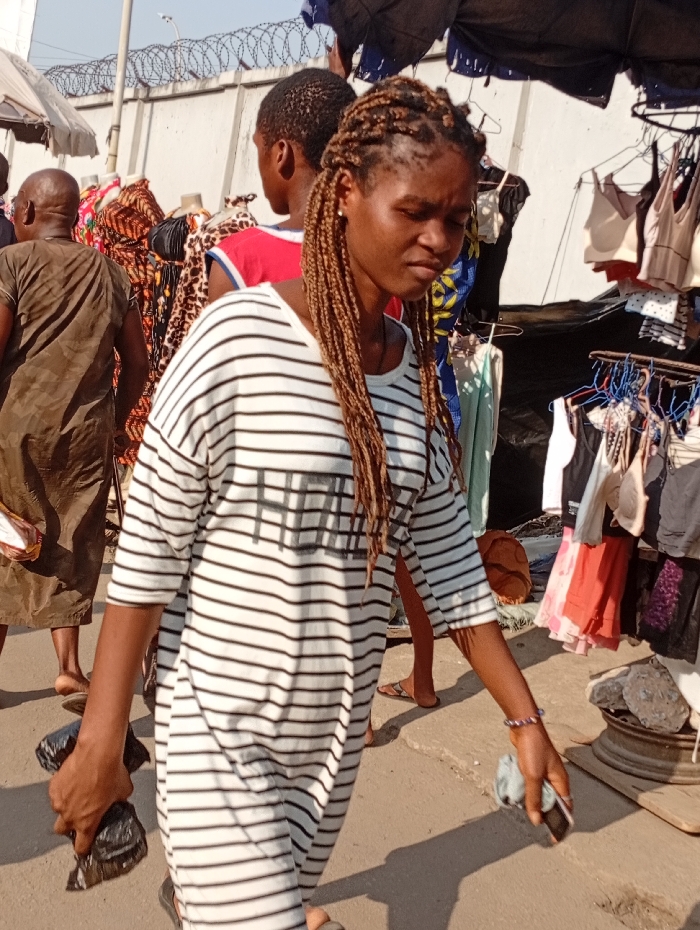 A woman with long, blonde braids wearing a striped black-and-white dress walks through an outdoor market. She holds a black plastic bag in one hand and a phone in the other. Clothing items hang on racks surrounding her, with other shoppers and vendors in the background. A wall with barbed wire sits at the top of the frame.