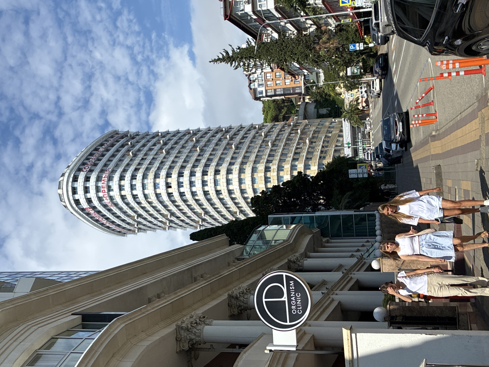This image shows a street scene in Sochi, Russia, with a tall, modern building dominating the skyline. The building, which appears to be a hotel or apartment complex, is curved and features numerous windows. Red lettering is visible on the upper portion of the building. To the left of the tall building, a more traditional building with columns and an awning is seen, featuring a circular sign that reads "ORGANISM CLINIC." Three young women are walking on the sidewalk in front of this building. One woman in a white top and beige pants is walking from left to right, with her hand near her forehead. Another woman in a white top and a light blue denim skirt is walking next to her, gesturing with her right hand. The third woman, in a white top and white shorts, walks to her right.
The street is paved and lined with parked cars, including a black SUV in the foreground. Orange and white traffic cones are placed along the curb. Further down the street, more buildings, trees, and a street sign are visible. The sky is blue with scattered white clouds, indicating a clear day. The overall impression is of a sunny day in a developed urban area.