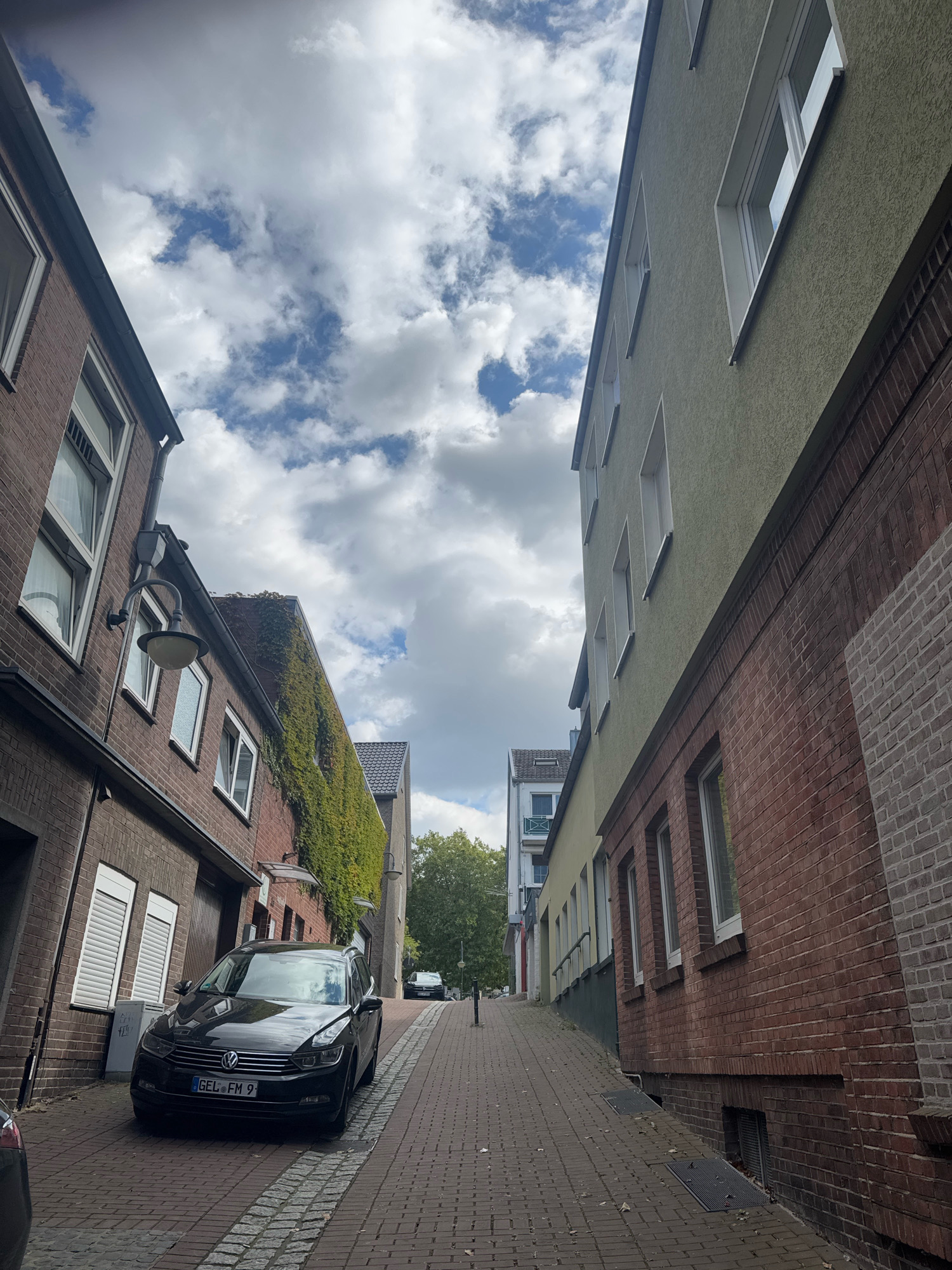 This is a low-angle, vertical photograph of a narrow street in Cleves, Germany. The street is paved with brick pavers and slopes upwards away from the viewer. On the left side of the street, there is a brick building with several windows and a downspout. A black Volkswagen sedan is parked parallel to the curb in front of this building. The license plate reads "GEL FM 9". A black car is partially visible on the left edge of the frame.
On the right side of the street, there is a building with a light green stucco facade above a red brick base. This building has multiple windows on its upper floors. Further down the street, additional buildings are visible, one with a yellow facade and a white upper section with balconies, and another with a grey tiled roof. Trees with green foliage are visible in the distance. The sky above is partly cloudy with large, white cumulus clouds against a blue sky. The scene appears to be one of a residential or commercial street in a European city.