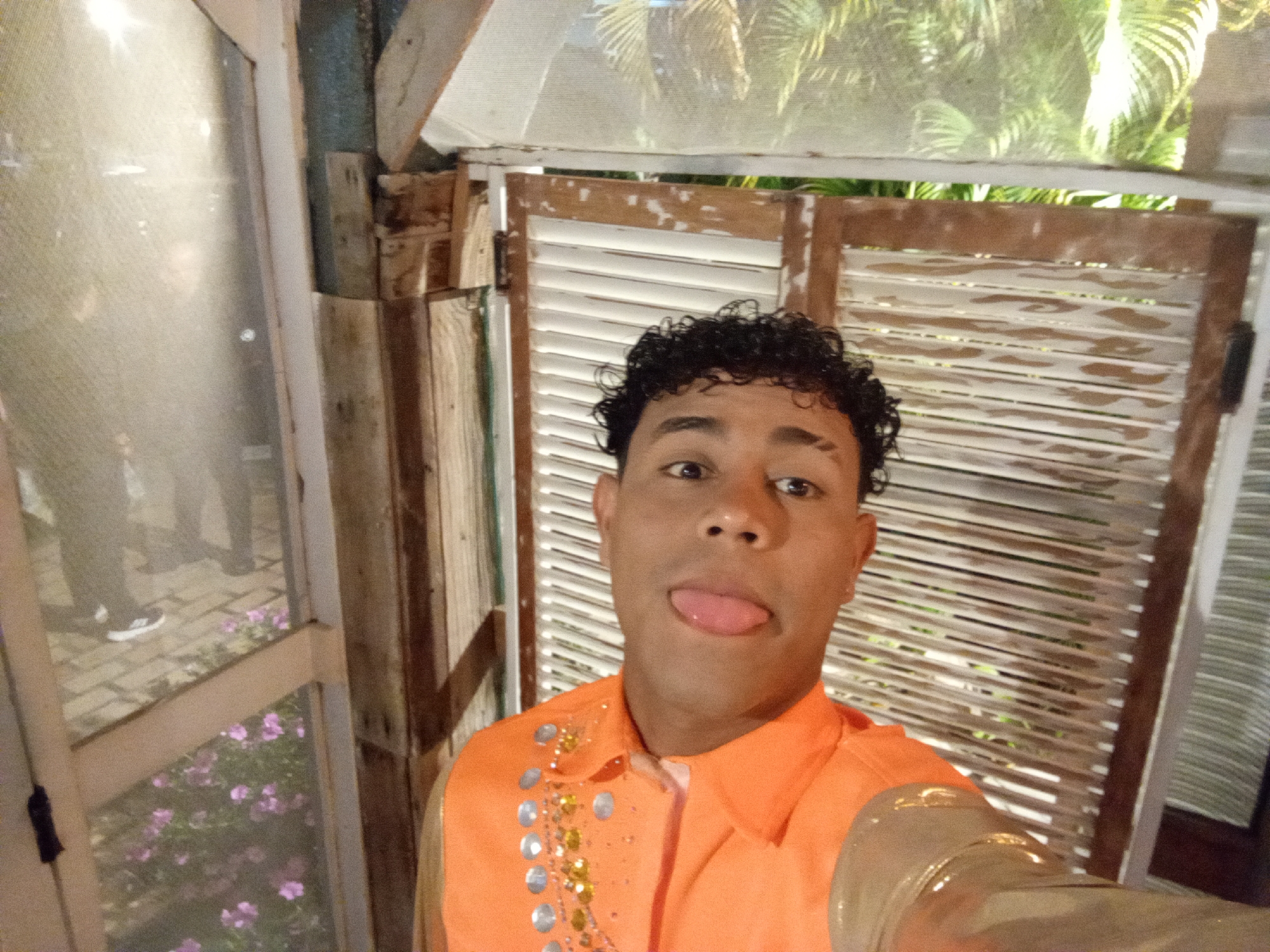 A person with dark curly hair, wearing an orange shirt with decorative embellishments, is taking a selfie. The person is sticking their tongue out playfully. The background consists of wooden structures, including shuttered doors and window frames. Through the window, there are purple flowers and legs of other people, possibly standing in a garden. Outside the shuttered doors, greenery including palm leaves is visible. The city of Porlamar, Venezuela is not directly verifiable from the image content.
