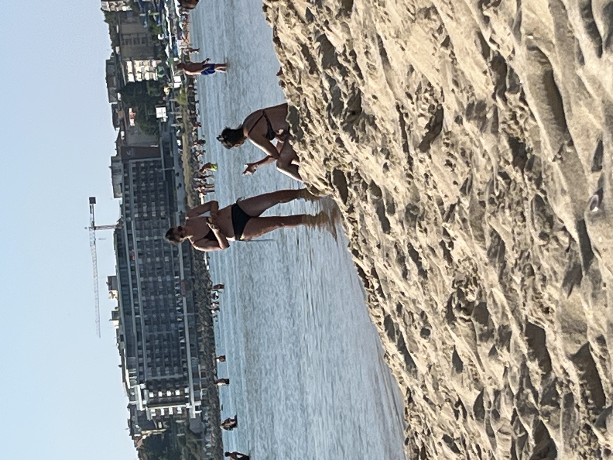 The image depicts a beach scene in Nesebar, Bulgaria, with the Black Sea in the foreground and a resort-style hotel with a construction crane in the background. Several individuals are visible in the shallow water and on the sand. In the foreground, two women are present; one stands in the water, holding an object, and the other sits on a sandy dune. Further out in the water, various people are wading and swimming. The sea is calm, and the sky is clear and bright. The sand in the foreground exhibits distinct rippled patterns from the receding tide.