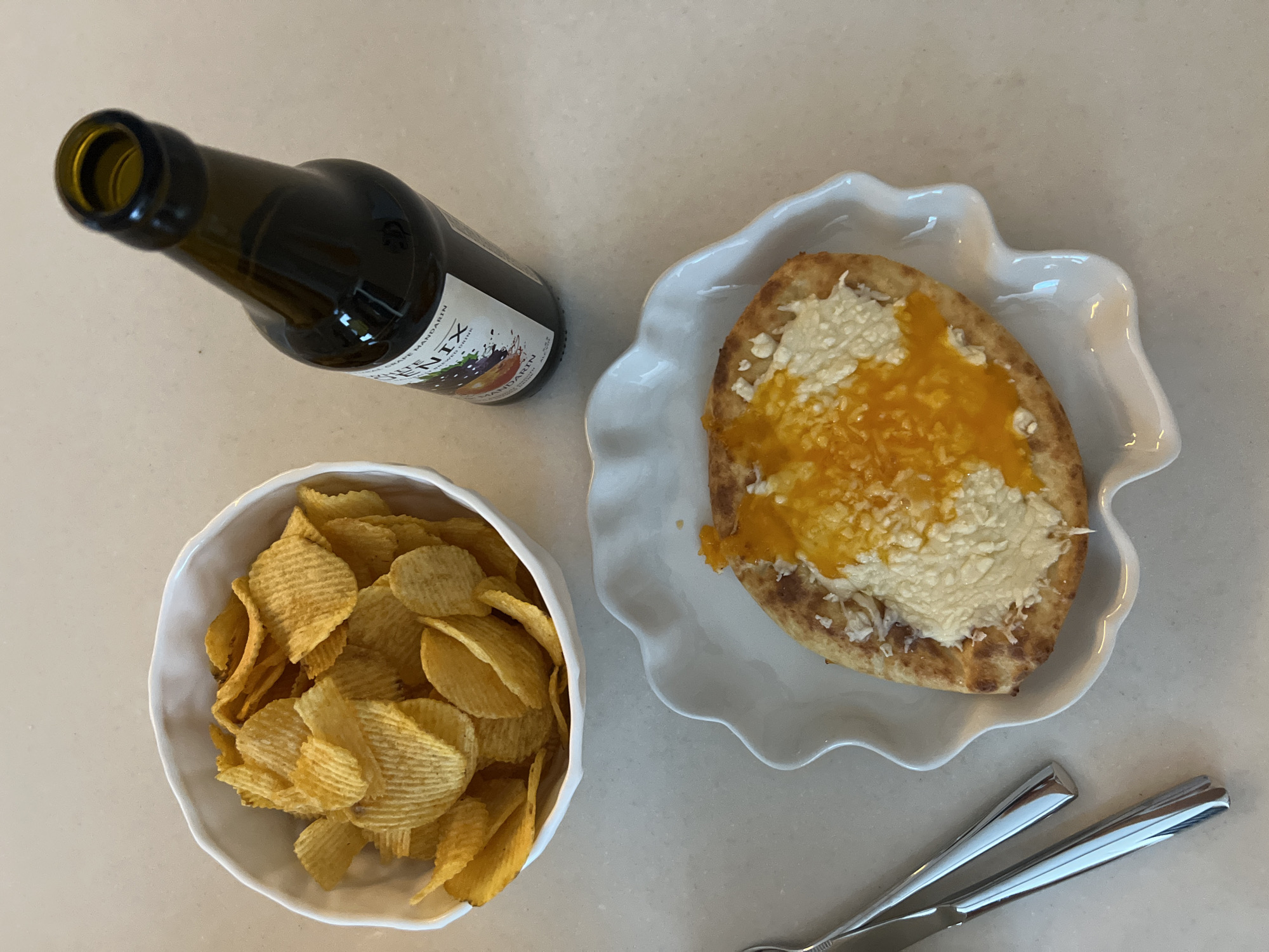 The image shows an overhead view of a light-colored countertop. There's a brown bottle with a label reading "QUENIX" and "Grape Mandarin" lying horizontally. Next to it is a white bowl filled with ruffled potato chips. A white plate with a wavy edge holds a piece of toasted bread topped with what appears to be orange jam and a white spread. A silver knife and fork are positioned near the plate. The location in the city of Новое, Russia is not identifiable from the image.