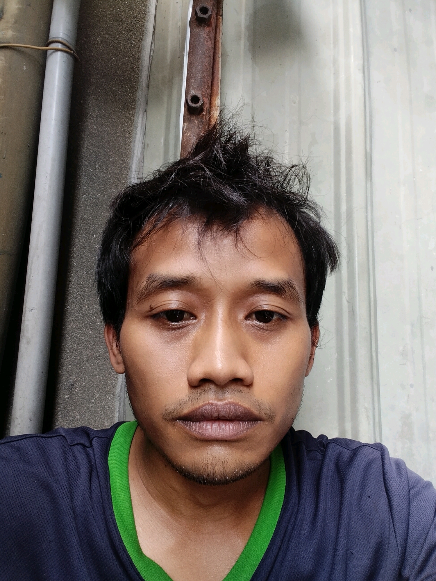 A close-up, head-and-shoulders selfie of a man in Douliu City, Taiwan. The man has dark, tousled hair, brown eyes, and light stubble on his upper lip and chin. He is wearing a dark blue t-shirt with a bright green collar. The background is a textured wall of corrugated metal, with a section of a gray pipe and a rusted metal beam to the left.