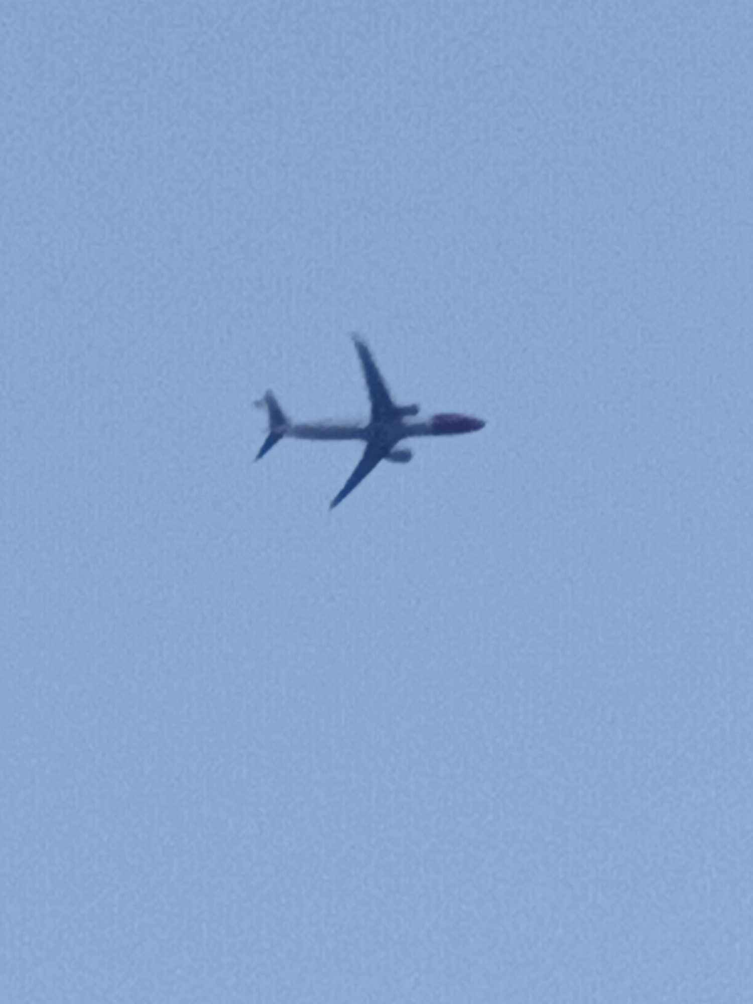 An airplane is seen from below, flying through a clear, light blue sky. The airplane is a dark silhouette against the bright sky, making it difficult to discern specific details. It appears to be a commercial passenger jet, with a long fuselage and wings extending outwards. The plane is angled slightly, suggesting it is in flight. The time of day appears to be daytime due to the bright sky, and the weather seems clear and fair with no visible clouds. The location is given as Prague, Czechia, but there are no visible landmarks or cues to confirm this. There is no discernible text in the image. The overall impression is a simple, clear shot of an airplane in the sky.