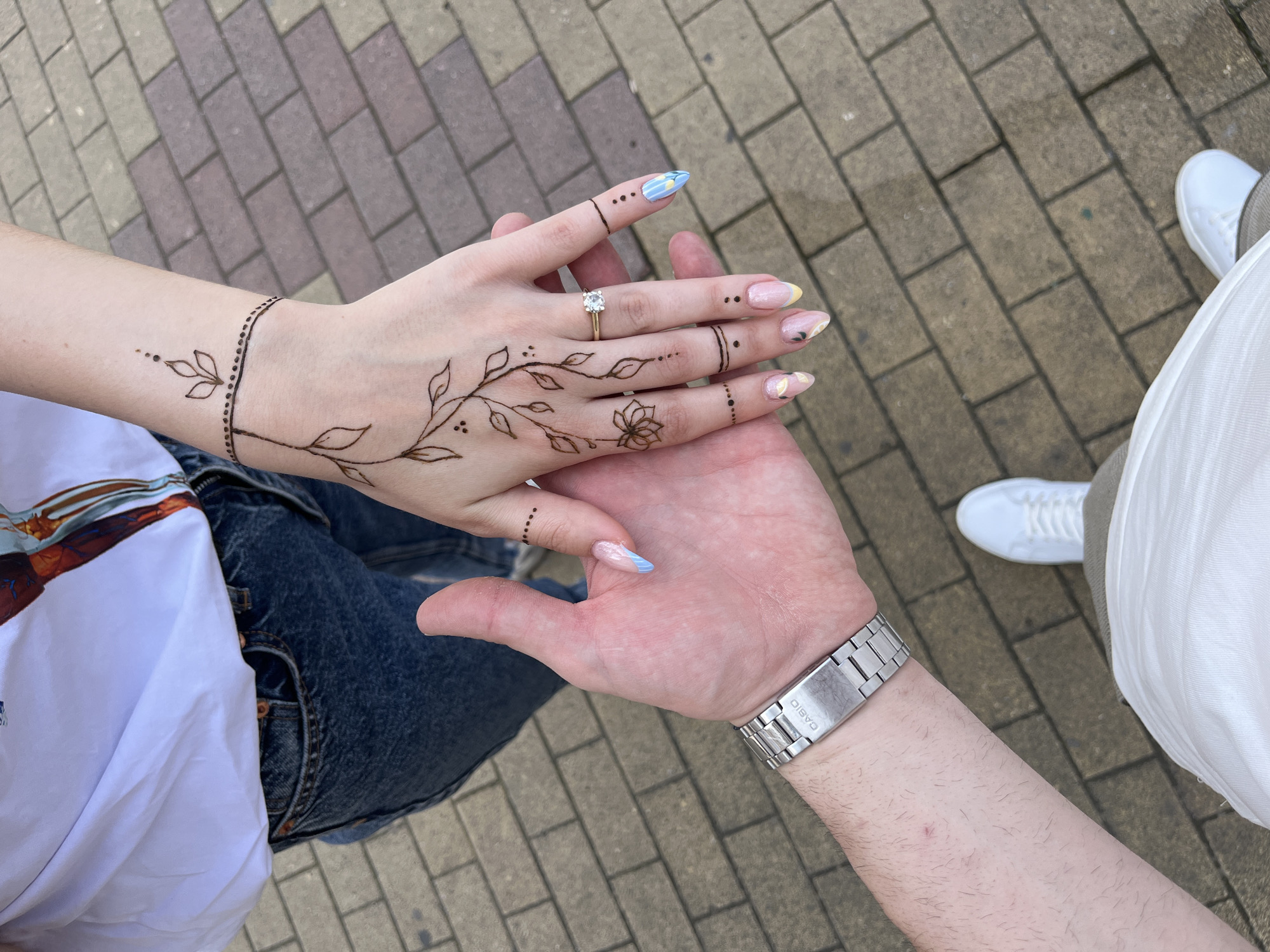 An outdoor, top-down shot in Madan, Bulgaria, depicts the interlocked hands of two individuals positioned above a paved surface composed of reddish-brown and gray rectangular pavers. The upper hand, belonging to an individual wearing a white graphic t-shirt and dark blue jeans, features intricate brown henna patterns including a leafy vine from the wrist to the fingers, a small flower on the ring finger, and dotted bands on multiple fingers. This hand also displays long, almond-shaped nails with varied pastel designs and a gold-toned ring with a clear solitaire stone on the ring finger. The lower hand, belonging to an individual wearing a white t-shirt, light brown pants, and white sneakers (partially visible at the bottom), exhibits a silver-toned Casio wristwatch on the left wrist. The visible forearm of the lower hand is lightly hairy.