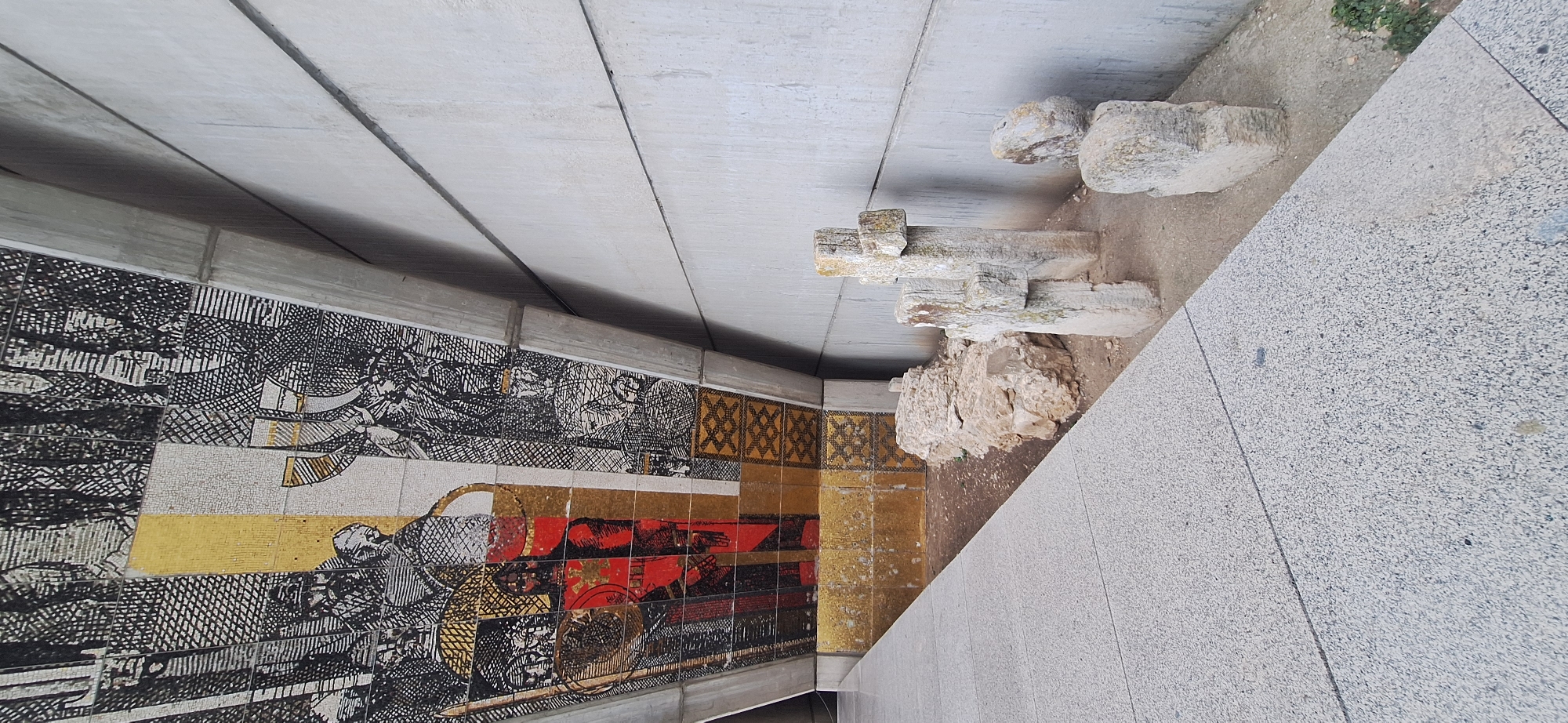 The image captures a perspective view of the Monument to the Bulgarian-Soviet Friendship in Shumen, Bulgaria. The foreground features a textured grey tiled walkway leading into the scene, angled to the left. To the right of the walkway, a patch of earth is visible, containing several weathered, light-colored stone objects. Among these are three upright stone structures resembling crosses, with one being taller and more intact than the others. There are also several rough-hewn stones scattered on the ground. In the background, a large, curved concrete structure forms the underside of the monument. This structure is adorned with a mosaic depicting figures, possibly historical or mythological, in black, white, yellow, and red tiles. The concrete wall to the right of the crosses is plain and textured. The lighting suggests it is daytime, with shadows indicating a light source from the left.