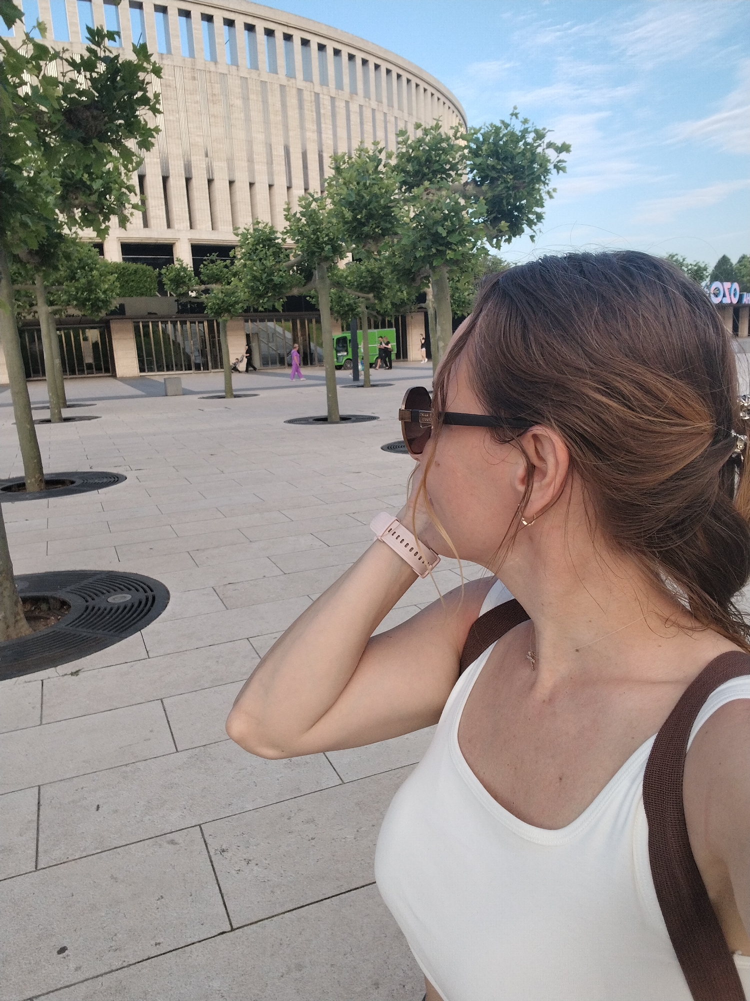 The image shows a woman from the chest up, facing to her right. She has brown hair tied back in a bun, is wearing sunglasses, a white tank top, a pink smartwatch, and a small necklace. She is standing in an outdoor plaza paved with light-colored rectangular tiles. In the background, there is a large, modern building with a curved facade and vertical elements. Trees are planted in circular grates in the plaza. The sky above is blue with some clouds. This appears to be the area in front of the "Expograd Yug" exhibition and convention center in Krasnodar, Russia, given the architecture of the building and the overall urban plaza setting.