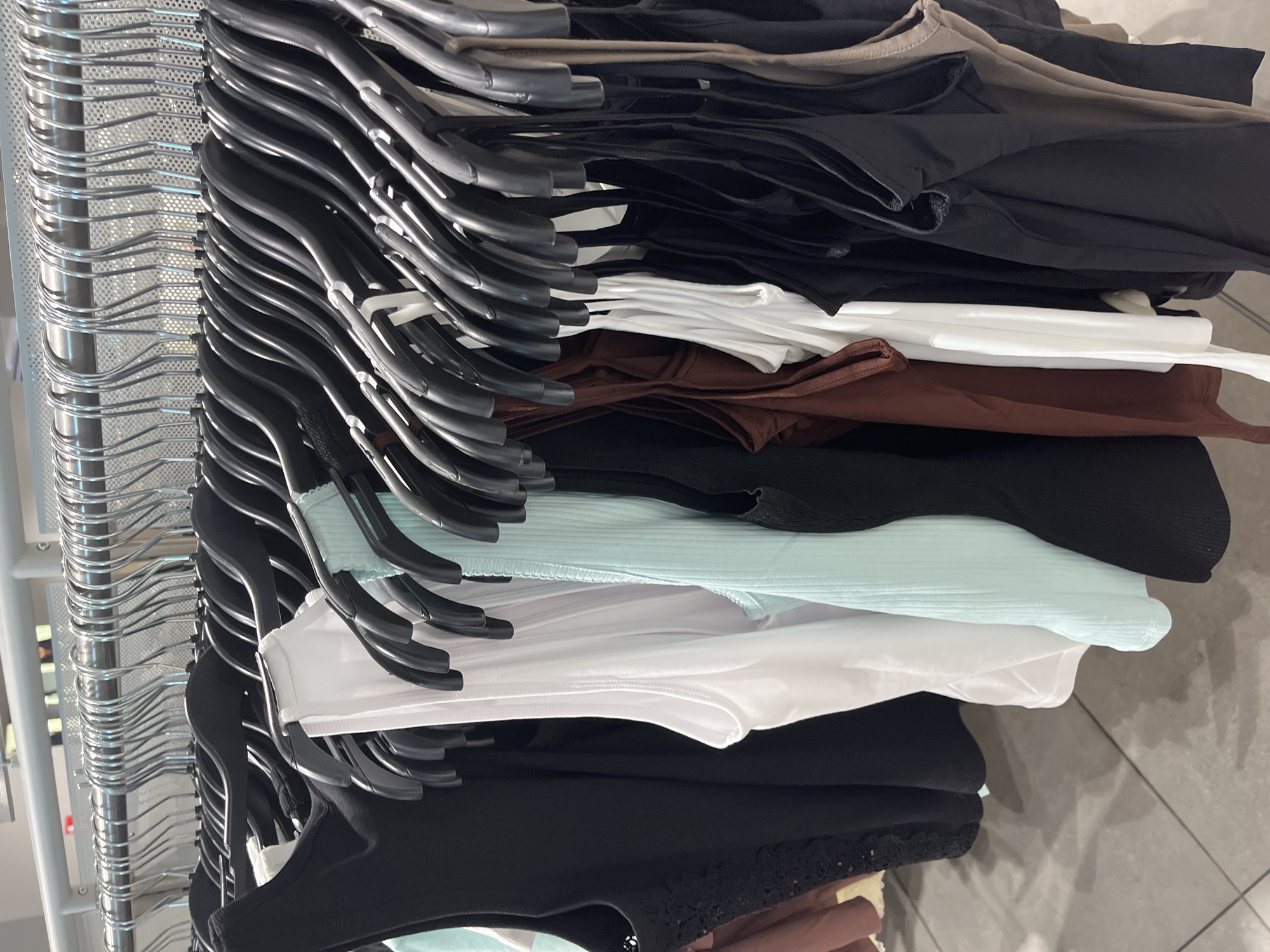 The image displays a close-up view of a clothing rack inside what appears to be a retail store in Blagoevgrad, Bulgaria. The primary subjects are numerous items of clothing and the hangers they are suspended from.
On the right side of the frame, a dense collection of garments, mostly tops or bodysuits, are neatly hung on black plastic hangers. These clothes are arranged in a stack, showcasing a variety of solid colors including black, white, various shades of brown (from dark to light beige), and a light blue or mint green. Some of the garments exhibit different textures, such as ribbed fabric on the light blue top and what appears to be lace detailing on a black top at the very bottom.
To the left, a metal clothing rail is visible, holding a long row of identical silver wire hangers. The rail itself has a perforated or mesh-like design. The overall activity depicted is the static display or storage of clothing. The lighting suggests an indoor setting, likely a commercial space. No people are visible, and there is no discernible text on any of the items or in the background. The floor beneath the rack is tiled, further indicating an indoor retail environment.