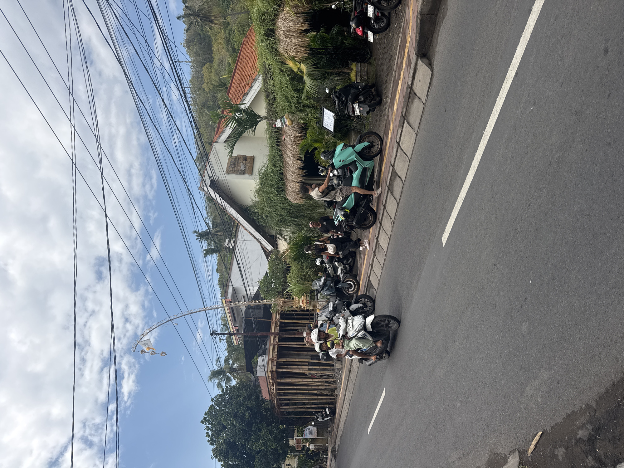The image depicts a street scene in Abang, Indonesia, featuring a road with a white lane marker. Several motorcycles are parked along the side of the road, and two individuals are riding a white scooter. In the background, a building with a thatched roof and lush green foliage is visible, along with trees and overhead power lines. The sky is partly cloudy. A bamboo structure is under construction to the left of the building.