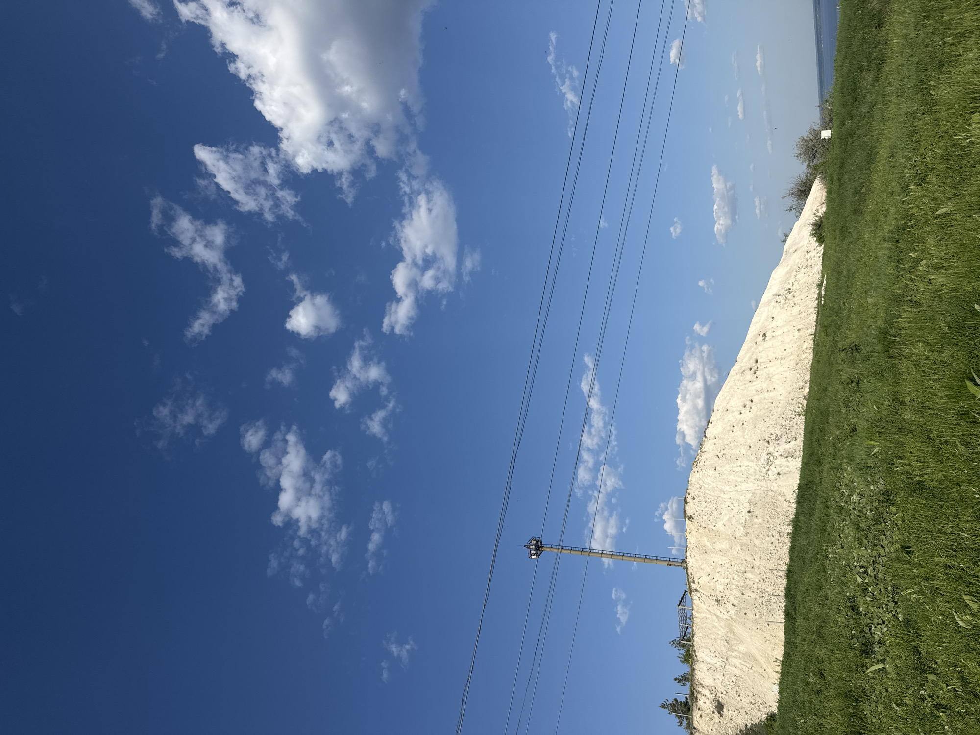 The image shows a landscape with a chalk cliff on the right side. Adjacent to the cliff is green grass. The sky is blue with scattered white clouds. Power lines extend across the sky, supported by a metal tower. The photograph was taken in the city of Khvalynsk, Russia.