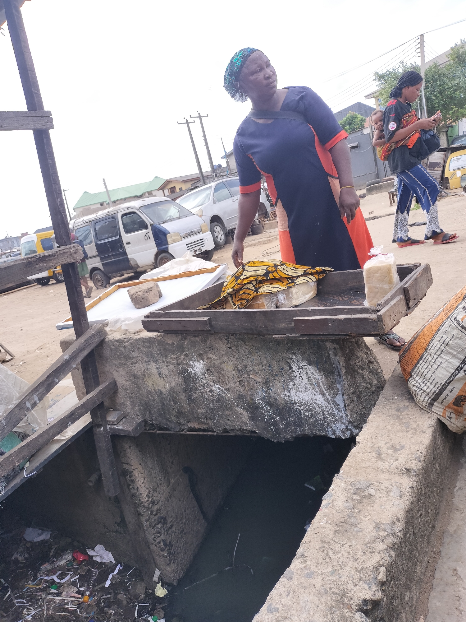 The scene is an outdoor market in Lagos, Nigeria. A woman wearing a blue and orange dress and a blue head covering stands behind a wooden tray on top of a concrete structure, where she is selling bread. The bread is wrapped in plastic. Next to the concrete is a bag of what looks like flour. The concrete structure also appears to be a drain with trash and water inside. In the background, there are other people walking by and some vehicles parked. The scene is likely located in a busy commercial area with various economic activities taking place. Another woman and a baby on her back pass by. Overhead powerlines can be seen in the background.