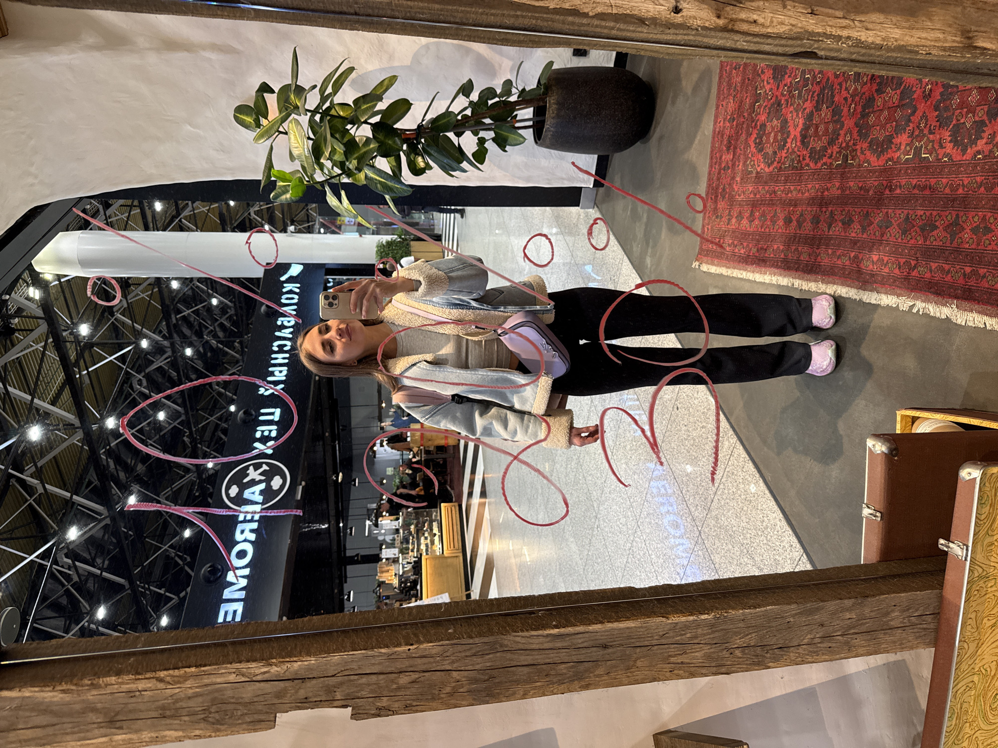 The image depicts a young adult female taking a selfie in a large mirror within a commercial complex in Khimki, Russia. The woman, with long brown hair, is wearing a light-colored top, a light blue denim-style jacket with a cream shearling lining, black pants, and light pink slip-on shoes. She holds a light-colored smartphone with three camera lenses in her right hand to capture her reflection. A small, light purple shoulder bag hangs from her left side.
The mirror, framed by thick, distressed wooden beams, displays handwritten red markings including an inverted "10," a percentage symbol, and other indistinct inverted numbers or squiggles, possibly "20" and "30." To the right of the woman's reflection, a large, dark green potted plant in a dark pot stands on a red oriental rug with intricate patterns. In the lower left foreground, parts of two decorative cases or storage boxes with metal clasps are visible.
The reflection in the mirror reveals a spacious indoor commercial environment with a high, industrial-style ceiling featuring exposed metal framework and numerous light fixtures. The floor is composed of light grey polished tiles. A tall, white vertical architectural column is visible in the mid-ground. In the deeper background reflections, various commercial counters or food stalls with display cases are present, along with a section of floor featuring black and white stripes. A prominent black sign with white, inverted Cyrillic text, reading "ЭМОЯХАХ ТЕХ" and including a circular logo, is reflected in the upper left background, indicating a specific store or area within the complex. Indistinct individuals are visible in the distant reflections.