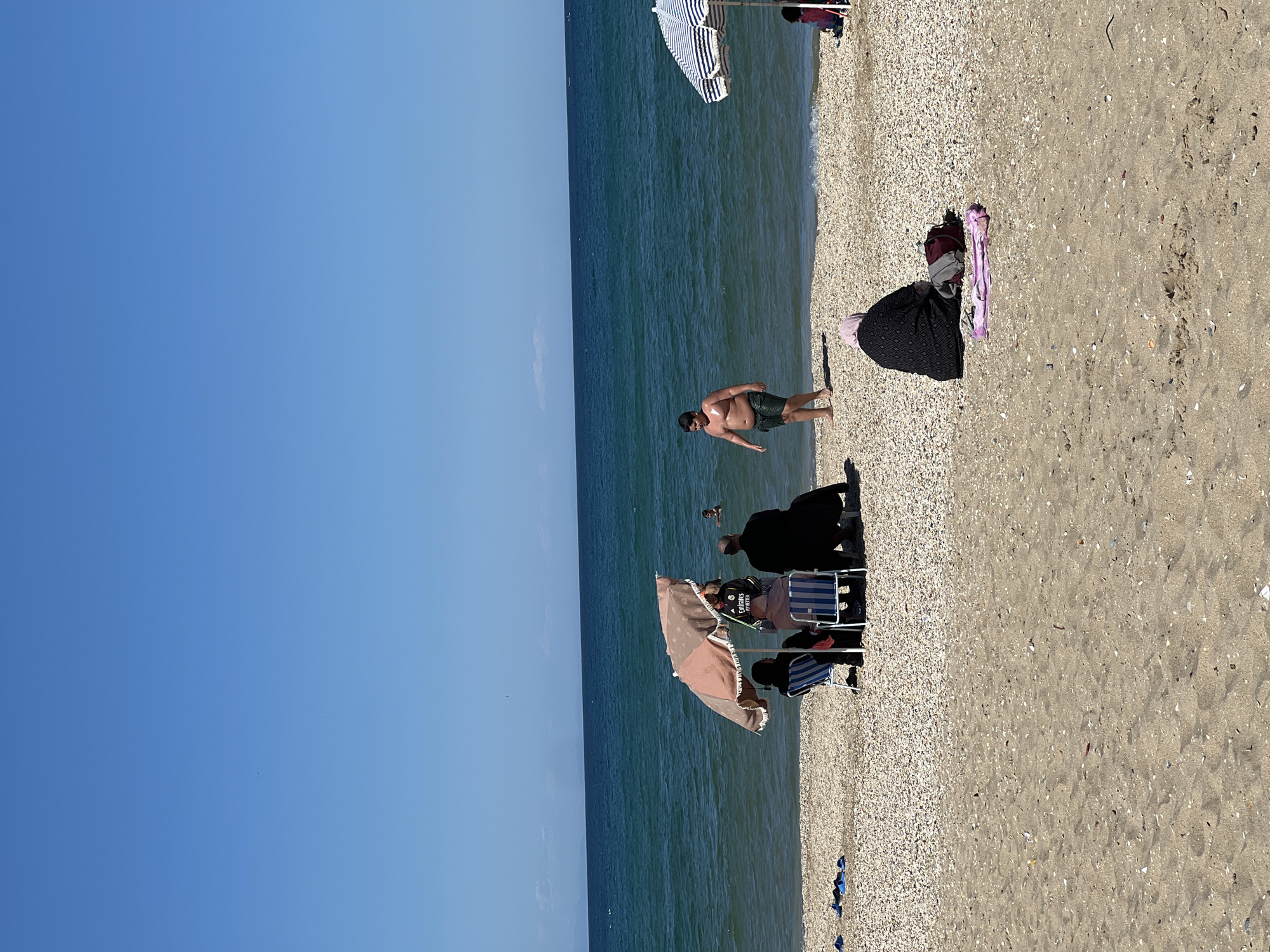 The image depicts a sunny beach scene in M'Diq, Morocco. The beach borders the sea and is occupied by several groups of people. A man is seen standing near the water's edge. Beach umbrellas and chairs are scattered across the beach, indicating a recreational environment. There are personal belongings of people as well scattered on the beach. The sky is clear blue, and the sea stretches to the horizon.
