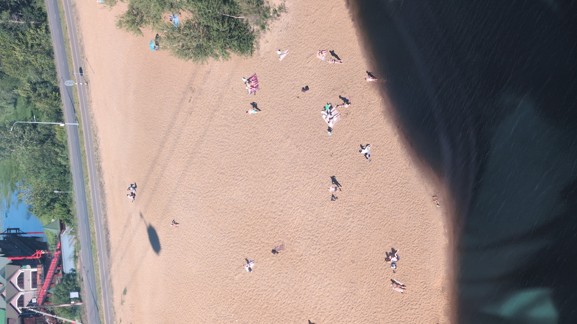 This aerial, high-angle shot captures a sunny day at a sandy beach in Nizhny Novgorod, Russia. Several people are scattered across the sand, engaged in various leisure activities. Some are sunbathing, lying on towels or directly on the sand, while others appear to be sitting or walking. A group of people are gathered together, possibly having a picnic or conversation. The beach is bordered by a road on one side, with lush green trees and vegetation. On the other side, the dark blue water of a lake or river stretches out, reflecting the bright sky. The scene suggests a relaxed atmosphere, with the clear, sunny weather indicating it's likely midday. There are no visible signs of text in the image. The composition emphasizes the expansive sandy area and the people enjoying the warm weather by the water.