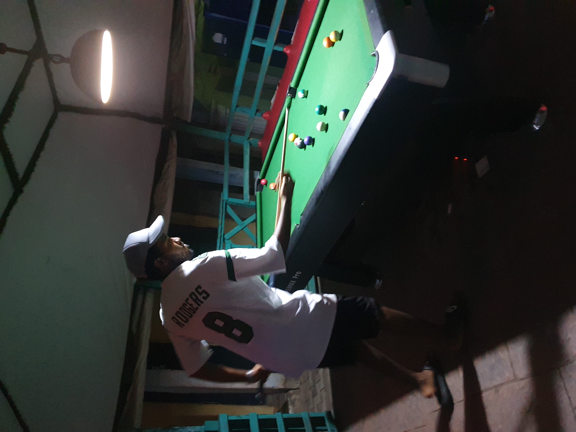 The image shows a man playing pool in an indoor setting. He is wearing a white jersey with the name "RODGERS" and the number "8" on the back, along with a baseball cap. The pool table is green and has several balls on it. The environment appears dimly lit, with a hanging light fixture overhead. Based on the request, the location is Lokoja, Nigeria, although no specific details in the image definitively confirm this.
