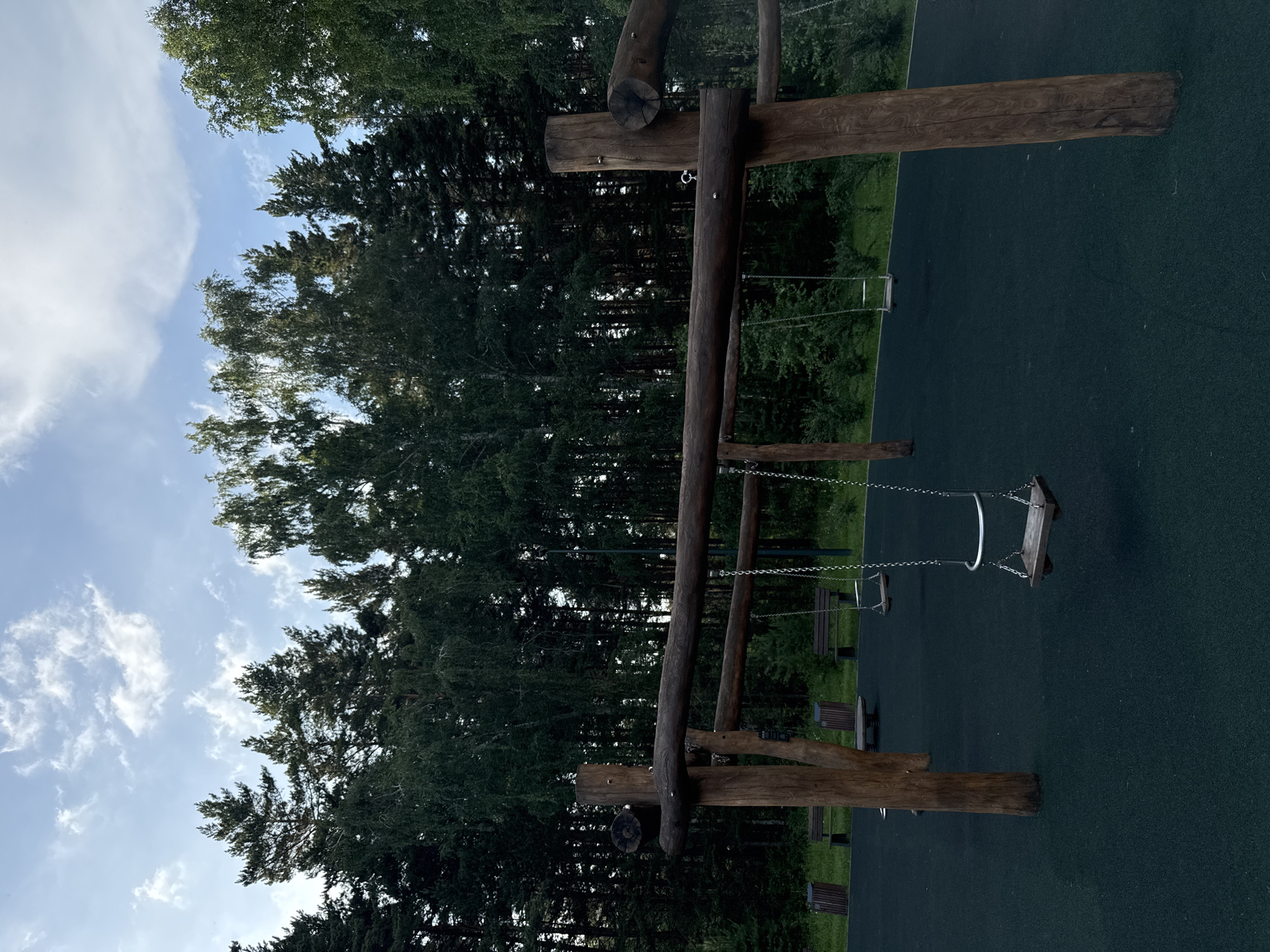 An outdoor playground with swings made of wood and chains is situated in front of a dense forest. The swings hang from a wooden frame supported by thick, rough-hewn logs. The ground beneath the swings is a dark, rubberized surface. In the background, several wooden benches and trash receptacles are visible, also near the edge of the forest. The sky above is partly cloudy with a blue hue. The image depicts a natural, serene environment, likely in a park or recreational area within the city of Krasnoyarsk, Russia. There are no people visible in the image.