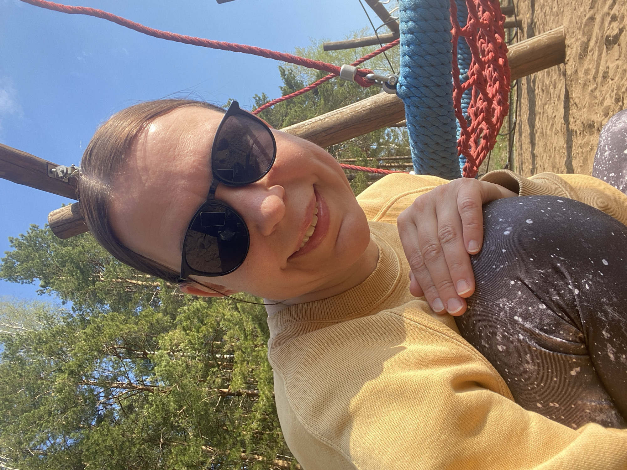 The image shows a smiling woman wearing sunglasses and a yellow sweater, sitting with her knees drawn up. She has light skin and brown hair. Her hand rests on her knees, which are covered in patterned leggings.
Behind the woman, a playground structure is visible, with wooden beams, a red rope, and a blue net. In the background, there are trees and a blue sky.
The location appears to be a playground near a wooded area, with possible indications of being in the city of Chaikovsky, Russia.
