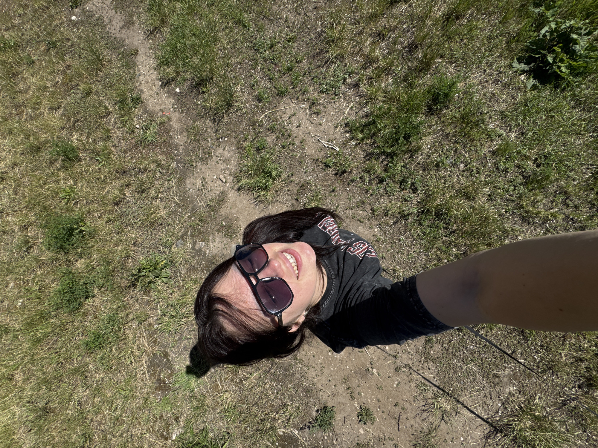 An overhead selfie captures a person outdoors, looking up towards the camera with a wide smile. The subject is a person with dark hair, wearing large, dark sunglasses perched on their head or forehead, and a black t-shirt with partially visible white or light-colored text, which appears to say "HARLEY" or a similar brand name. Their right arm, holding the camera, is visible extending into the frame from the right side.
The setting is an open, unpaved area under bright daylight, suggesting a sunny day, as indicated by clear shadows cast by the person and vegetation. The ground beneath is a mix of dry, patchy green and brown grass, interspersed with areas of exposed dirt and small stones or debris. The overall impression is an outdoor, informal environment like a field or a park. The person's joyful expression and the bright lighting convey a positive and casual mood.