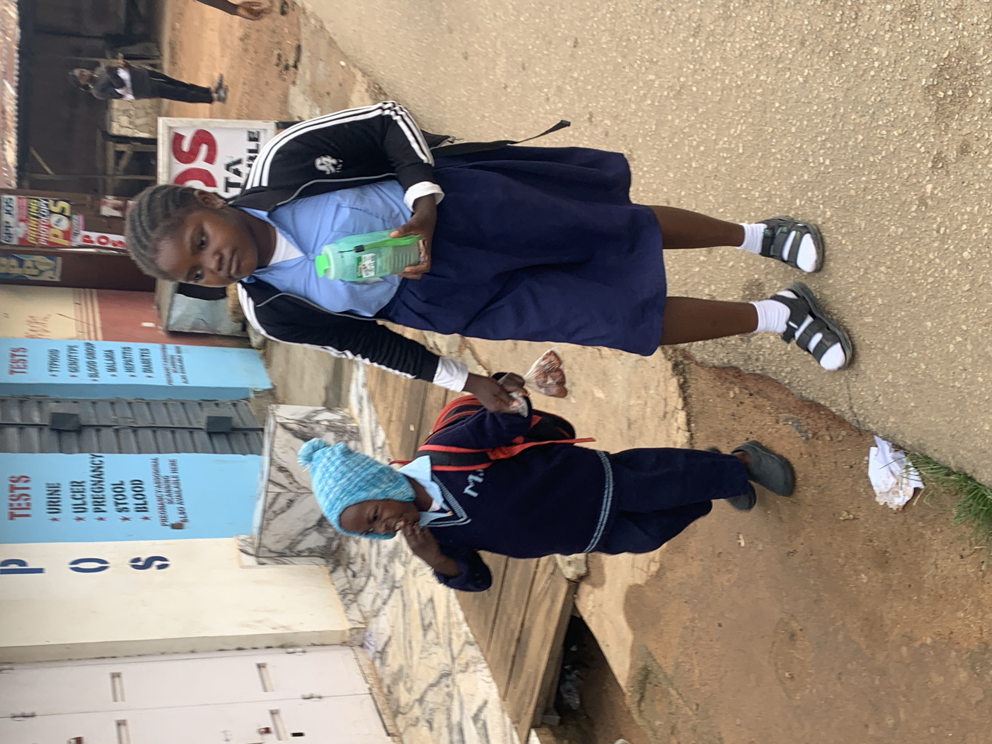 This media file captures a scene of two young girls, likely students, in what appears to be a street in Jos, Nigeria. The image is oriented vertically, with the ground taking up the lower portion and the subjects and background in the upper.
The first girl, positioned higher in the frame, is wearing a blue school uniform shirt and a dark blue skirt. She has dark braided hair and is holding a translucent green water bottle in her right hand. She is wearing dark sandals with white socks. Behind her, a sign with large red letters "POS" is visible, as well as other text related to medical tests like "URINE", "ULCER", "PREGNANCY", "STOOL", and "BLOOD GROUP".
The second girl, slightly lower and to the right, is wearing a similar dark blue school uniform, including a v-neck sweater. She has on a bright blue knitted beanie and dark shoes. She appears to be smiling. This girl also has a backpack on.
The background shows a street with a textured pavement. There are buildings and signs, including one that says "TA LE" in large letters. The overall impression is a daytime scene, possibly after school or during a break, in an urban or semi-urban setting in Nigeria. The presence of medical test signs suggests proximity to a clinic or laboratory. The girls' attire indicates they are likely attending school.