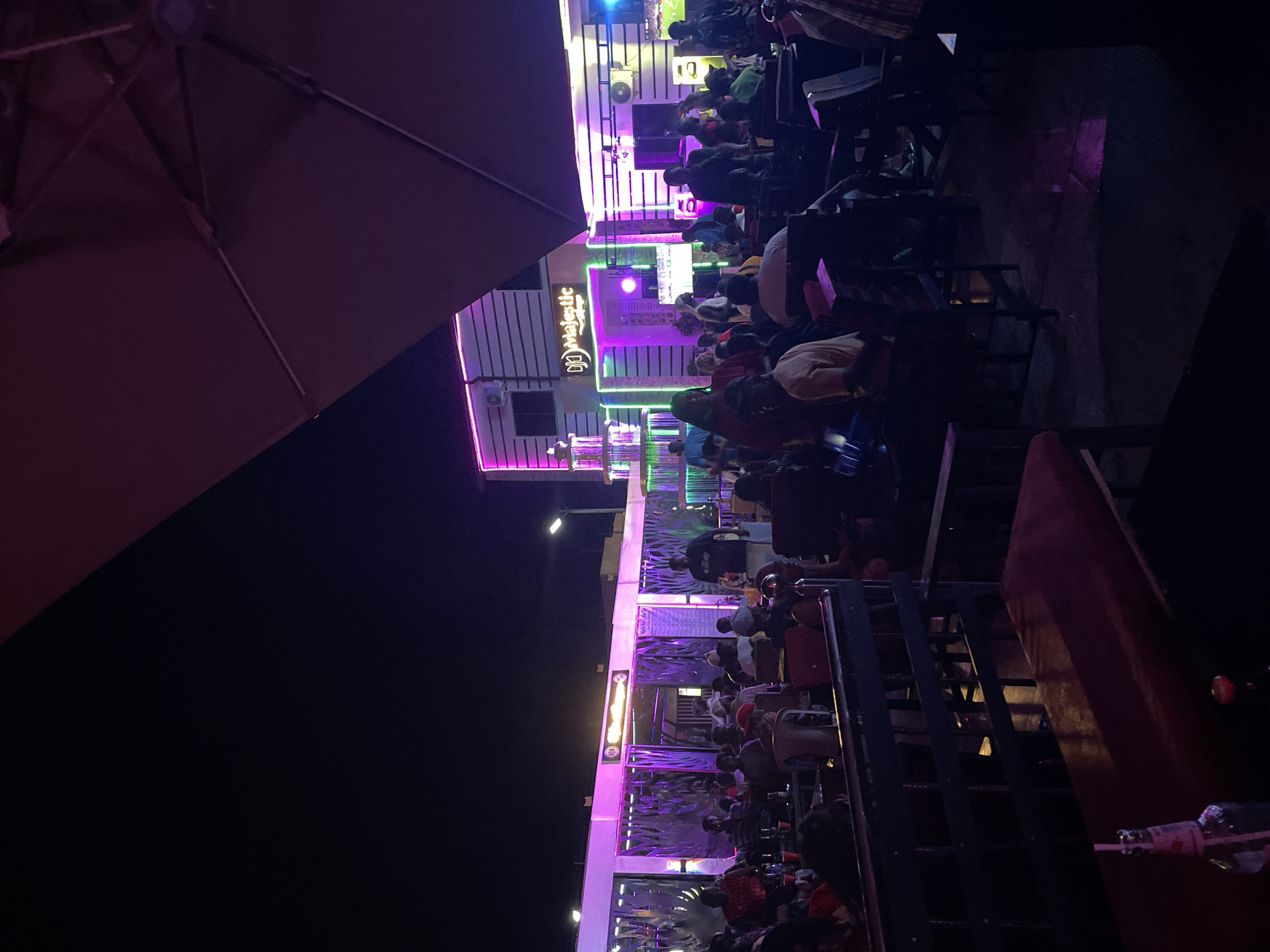 The scene depicts an outdoor gathering at the "Majestic" establishment in Tumfure, Nigeria, illuminated by vibrant purple and green neon lighting. Numerous people are seated at tables and chairs, suggesting a social event or dining experience. An umbrella covers the tables, while the establishment itself is a multi-story building with distinct architectural features.