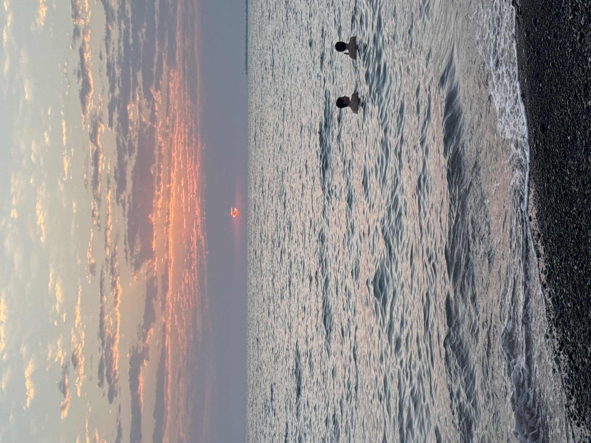 Two individuals are swimming in the sea at sunset in Gulrypsh, Abkhazia. The sun, partially obscured by clouds, casts a warm, pinkish-orange glow across the sky. The sea is calm with gentle waves lapping at a pebble beach in the foreground. The sky is filled with scattered clouds, some tinged with the colors of the sunset. The water appears a muted blue-grey, reflecting the ambient light. Both individuals appear to be young, with only their upper bodies visible above the water. They are positioned a moderate distance from each other. The overall scene conveys a sense of tranquility and natural beauty.