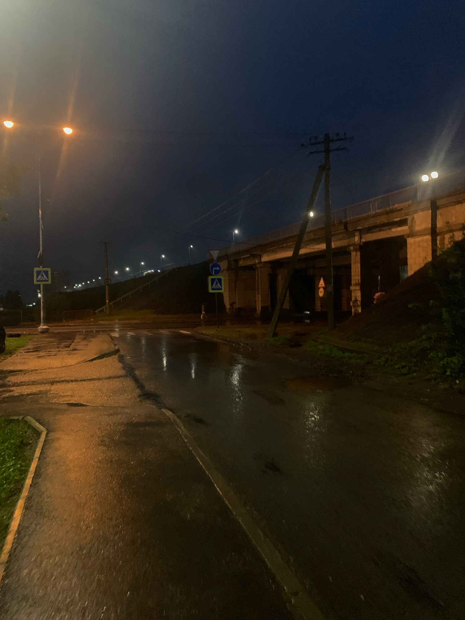 The image depicts a street scene at night in Arkhangelsk, Russia, likely in a suburban or industrial area. The asphalt road is wet, reflecting the light from streetlamps and the underpasses of an elevated roadway. There are several streetlights visible, illuminating the scene with a warm, orange glow. A pedestrian crossing sign is present on the left side of the road. Further ahead, a pedestrian overpass or bridge stretches across the scene, supported by concrete pillars. Several traffic signs are visible, including a speed limit sign of 40 and a blue directional sign with an arrow. The sky is dark, suggesting late evening or early morning. No individuals are clearly visible in the frame, although a shadowy figure may be present near the right side of the overpass. The overall atmosphere is damp and somber, consistent with recent rain.