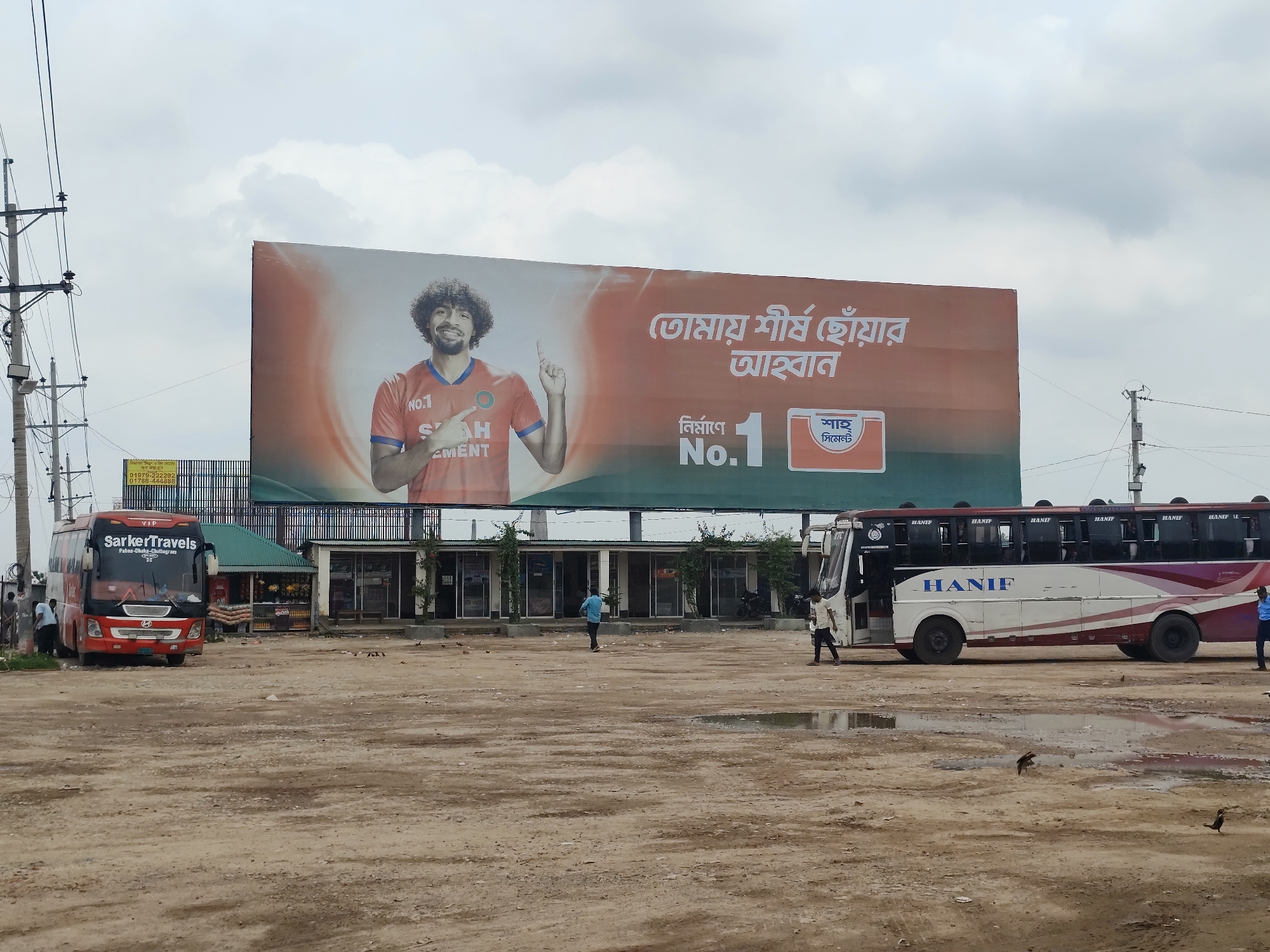 The image shows a bus station in Hatikumrul, Bangladesh. A large billboard advertising "Shah Cement" dominates the background, featuring a man in a sports jersey. In the foreground are two buses, one branded "Sarker Travels" and the other "Hanif." Several individuals are standing around the buses and in the open dirt lot. A one-story building runs along the base of the billboard. Utility poles are visible on the left side of the image. The ground is unpaved and dusty, with puddles of water. The sky is overcast.