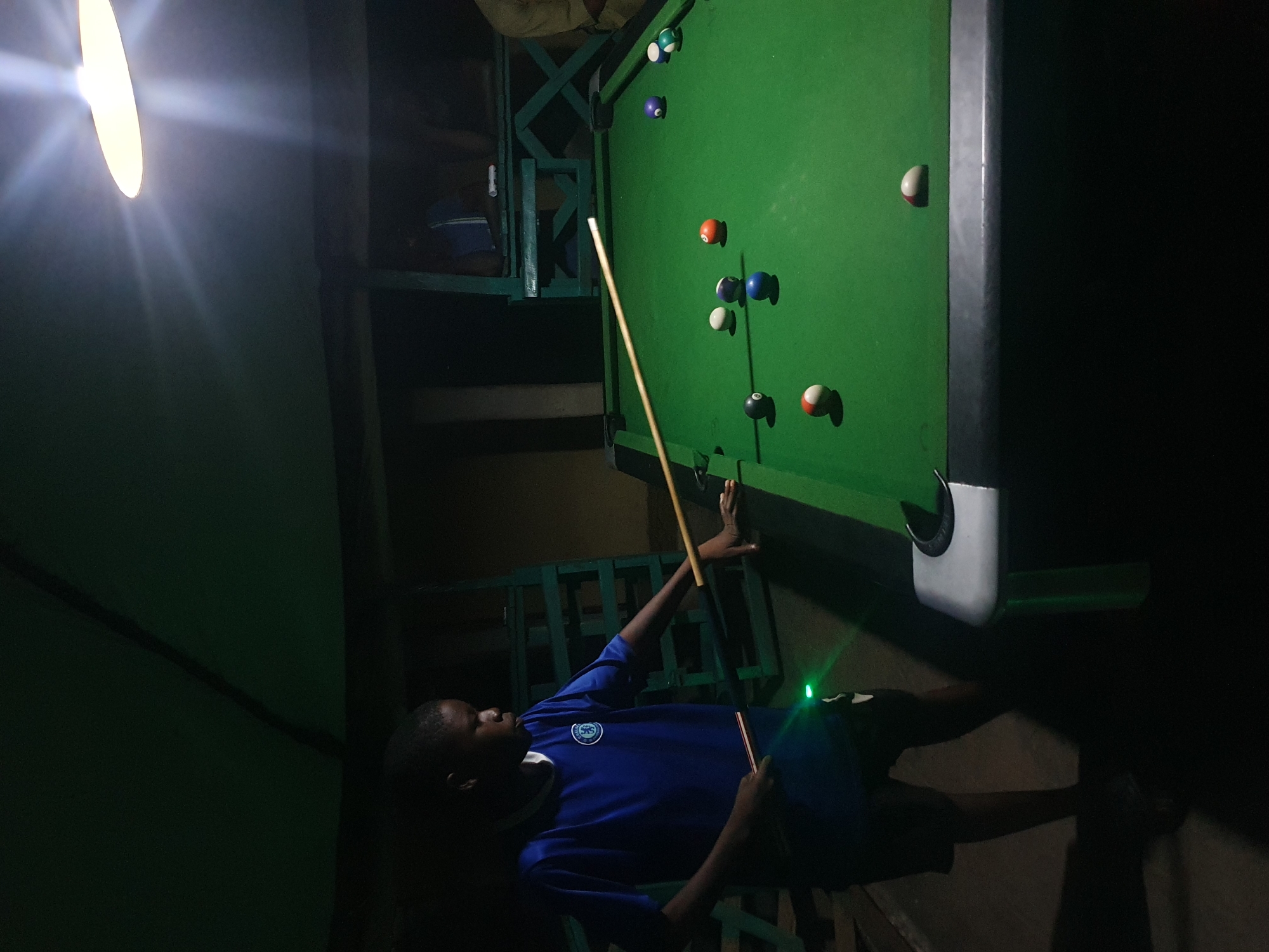 A young Black boy, wearing a blue t-shirt with a white trim around the collar and a crest on the left breast, is playing billiards. He is holding a cue stick and appears to be aiming for a shot on the green felt-covered table. Several billiard balls are scattered across the table, including a white cue ball, a red ball, a blue ball, and other striped and solid colored balls.
The setting appears to be an indoor, dimly lit space, possibly a recreational area or a backyard setup. A bright, possibly fluorescent, light fixture is visible on the left side of the image, casting a strong glare and illuminating part of the scene. The background is dark and somewhat indistinct, with hints of green metal furniture or shelving. The overall atmosphere suggests a casual, possibly evening, activity. The boy's posture and focus indicate he is engaged in the game. There is no visible text in the image. The location context provided is Lokoja, Nigeria.