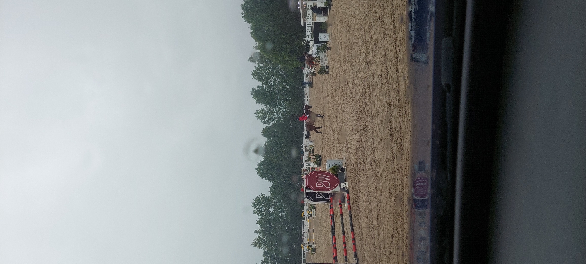 The image shows an outdoor equestrian event, likely a show jumping competition, on a cloudy day. The ground is covered in sand, and several jumps are set up. In the middle ground, a rider in a red jacket is on a brown horse, approaching a jump. To the right, another horse and rider are visible. In the background, there is a line of trees and white fencing. A reflection of the event is visible on a dark, glossy surface in the foreground, suggesting the photo was taken from inside a vehicle. The logo "BW" is visible on a banner. The location is identified as Ziniūnai, Lithuania.