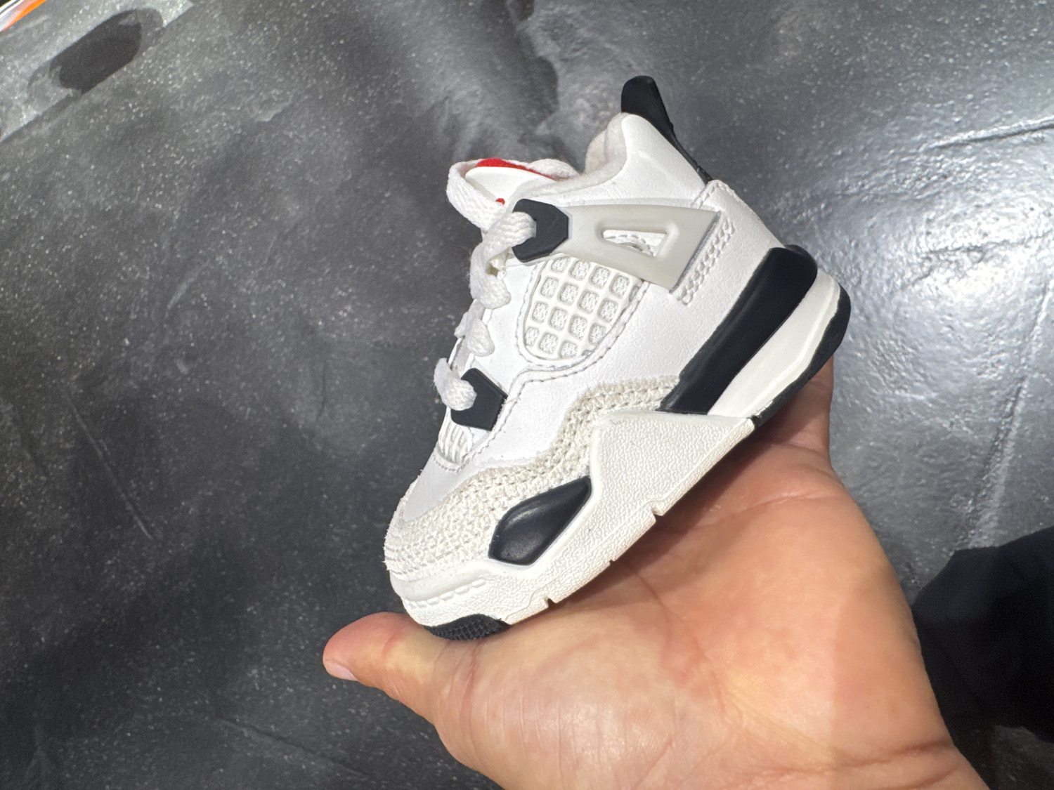 A hand, possibly an adult's, holds up a single, miniature sneaker against a dark, speckled, and somewhat textured background. The small shoe is predominantly white with black and subtle red accents, resembling a baby or toddler-sized Air Jordan 4.
The sneaker features white laces tied at the front. Its upper is made of white material, likely synthetic leather, with a distinctive grid-like pattern visible on the side panel near the laces. Black elements are incorporated into the eyelets and parts of the midsole, providing contrast. A light grey or beige textured material, possibly suede or a similar fabric, wraps around the toe area and extends along the lower side of the shoe. A small hint of red can be seen on the inner lining or tongue of the shoe, peeking out from the top. The sole is thick and white, with black sections on the bottom and sides.
The background appears to be a concrete or tiled surface, dark grey with lighter flecks and patches, suggesting a well-worn or industrial floor. The lighting seems even, not indicating a specific time of day. The hand is positioned to showcase the shoe, which is angled slightly upwards and to the right, highlighting its design and small size. There is no visible text in the image. Given the context, this scene is set in Barcelona, Spain.