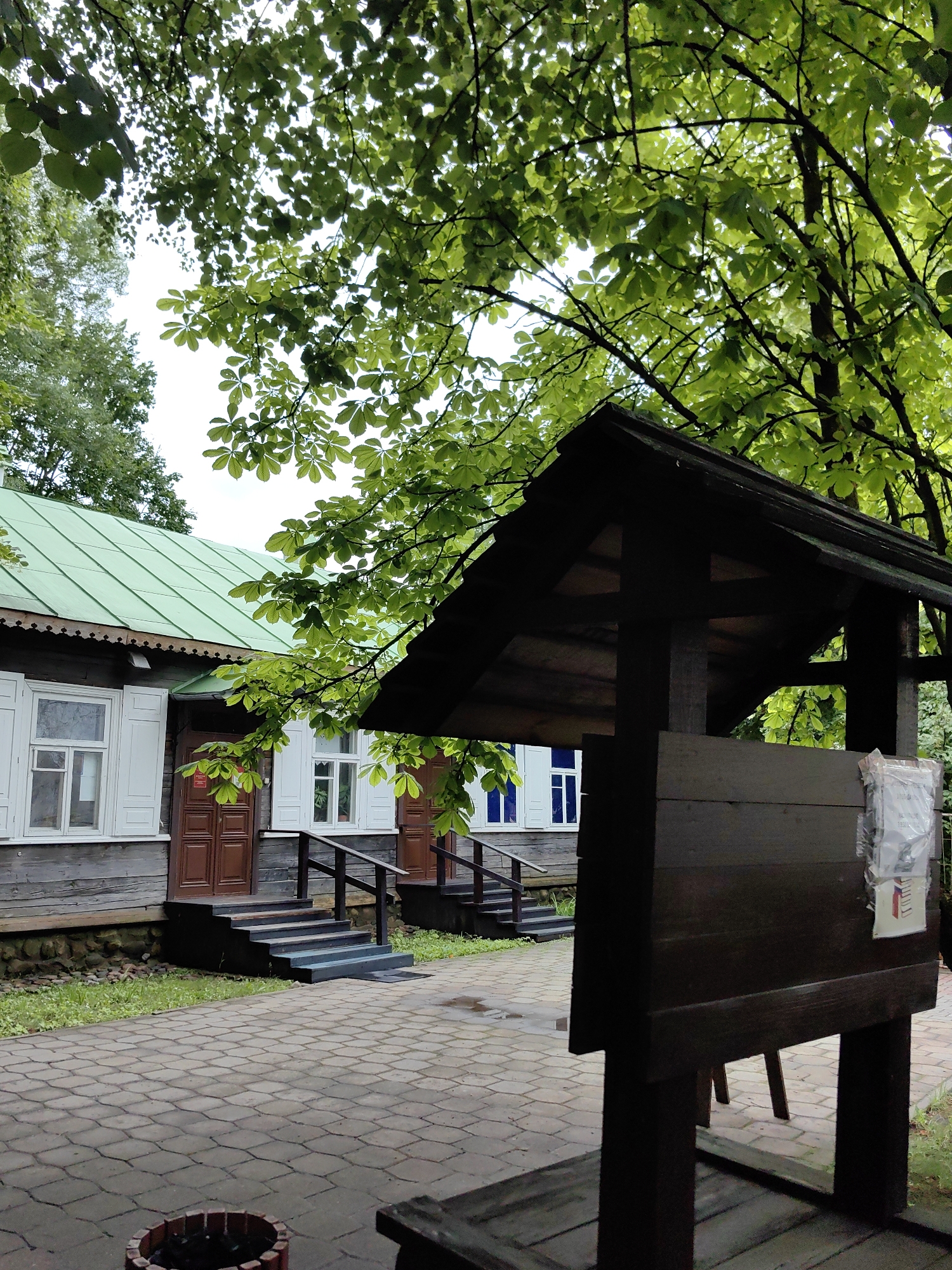 In Minsk, Belarus, a rustic wooden building with a green metal roof stands partially obscured by lush green foliage. The building features several windows with white shutters and brown wooden doors leading up to it via dark wooden stairs. In the foreground, a dark wooden structure with a sloped roof stands on a paved courtyard. The paving stones are arranged in a regular, repeating pattern. The environment is verdant, with trees and grass surrounding the building. The sky is overcast.