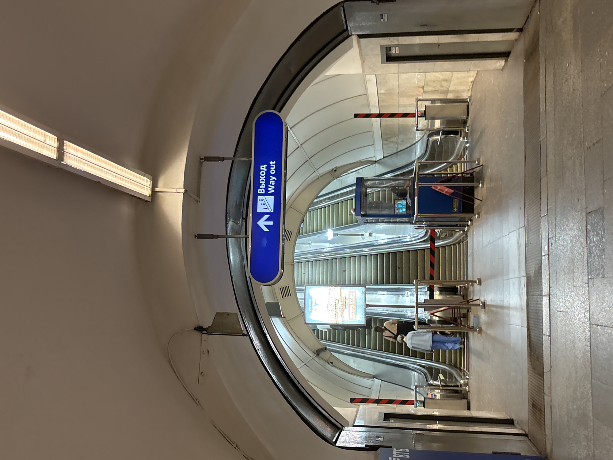 The image shows an interior view of a metro station in Saint Petersburg, Russia. Two escalators ascend into the distance, with a blue sign above them reading "Выход Way out" in Russian and English, indicating the exit. Several people are visible, including two individuals walking up one of the escalators. The architecture features a curved, white ceiling with a long fluorescent light fixture. Barriers with red and black tape cordon off areas near the escalators. A blue booth-like structure is positioned next to the escalators, and a poster is displayed on a stand in front of the ascending escalators. The floor is tiled, and a metal grate is visible in the foreground.