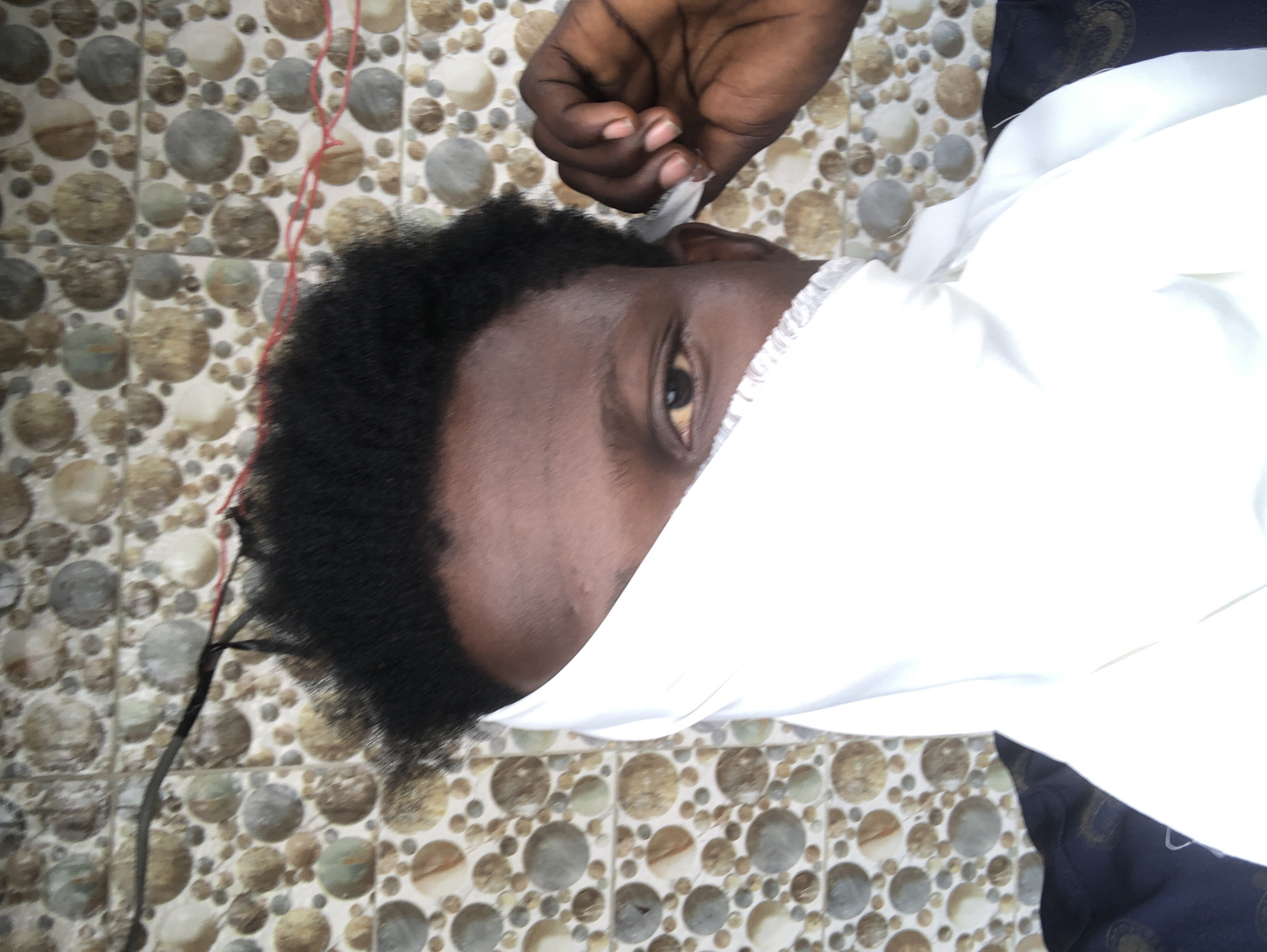 A close-up, waist-up shot captures a person's face partially obscured by a white fabric. The person has dark skin and black, short, kinky hair. Their right eye is visible and brown. Their left hand is raised to their face, adjusting the white fabric. The background features a wall with a circular, stone-like pattern in shades of brown, beige, and gray. Black and red electrical wires are visible against the wall. The lower portion of the image shows the person wearing a dark blue garment with a gold pattern. The scene appears to be indoors or in a covered outdoor area, possibly a home or shop in Ajah, Nigeria, indicated by the tiled wall and visible electrical wiring.