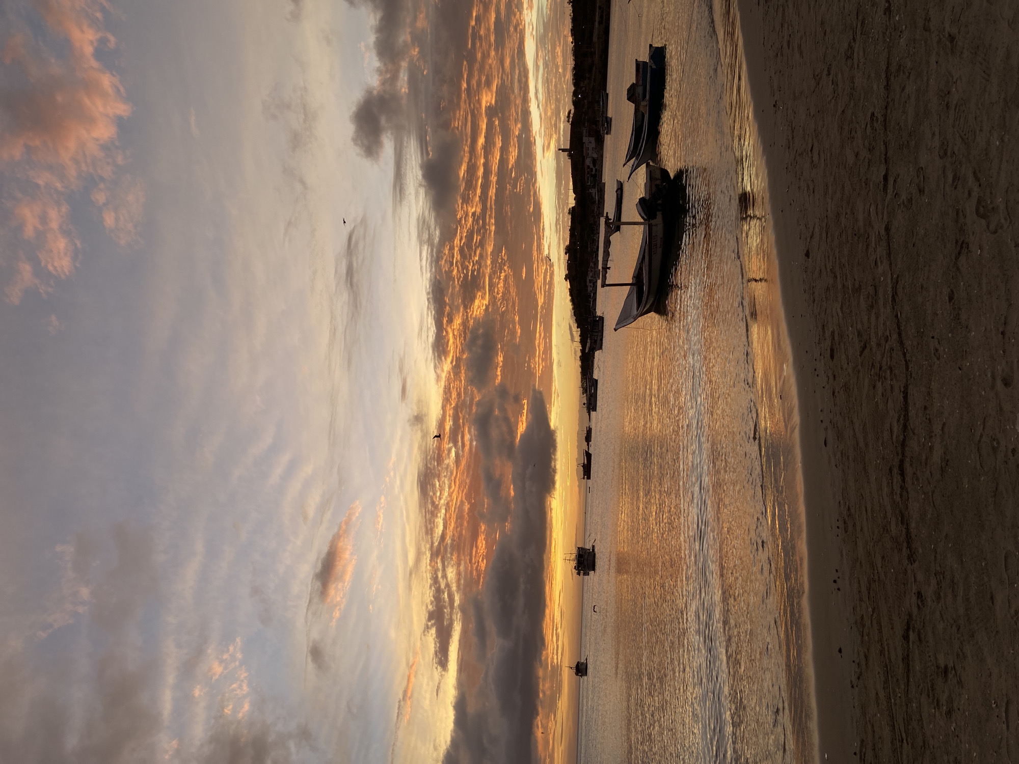 The image depicts a serene sunset over the bay of Juan Griego, Venezuela. The sky is ablaze with hues of orange, pink, and yellow, painted across a canvas of scattered clouds. The tranquil water reflects the vibrant sky, creating a mirror-like surface. Several small boats are anchored in the bay, their silhouettes stark against the colorful backdrop. In the foreground, a sandy beach stretches along the shoreline, with gentle waves lapping at the sand. The scene is peaceful and evokes a sense of calm and natural beauty.