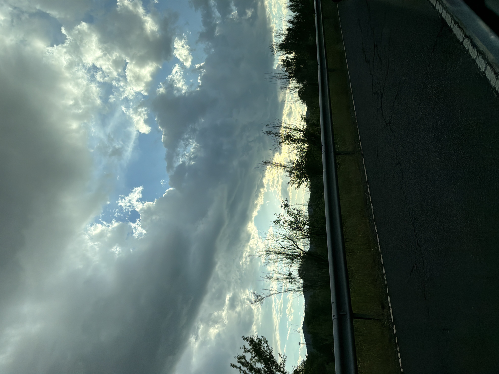 The image captures a view of a dramatic sky with billowing clouds, some obscuring the bright patches of blue sky. A dark, asphalt road stretches along the right side of the frame, bordered by a metal guardrail and green vegetation. A line of trees and a distant hill appear on the horizon, silhouetted against the sky. While the image does not provide enough information to confirm the exact location in Kirkovo, Bulgaria, the presence of a road, vegetation, and distant hills is consistent with a landscape potentially found in that region. There are no people visible in the image.