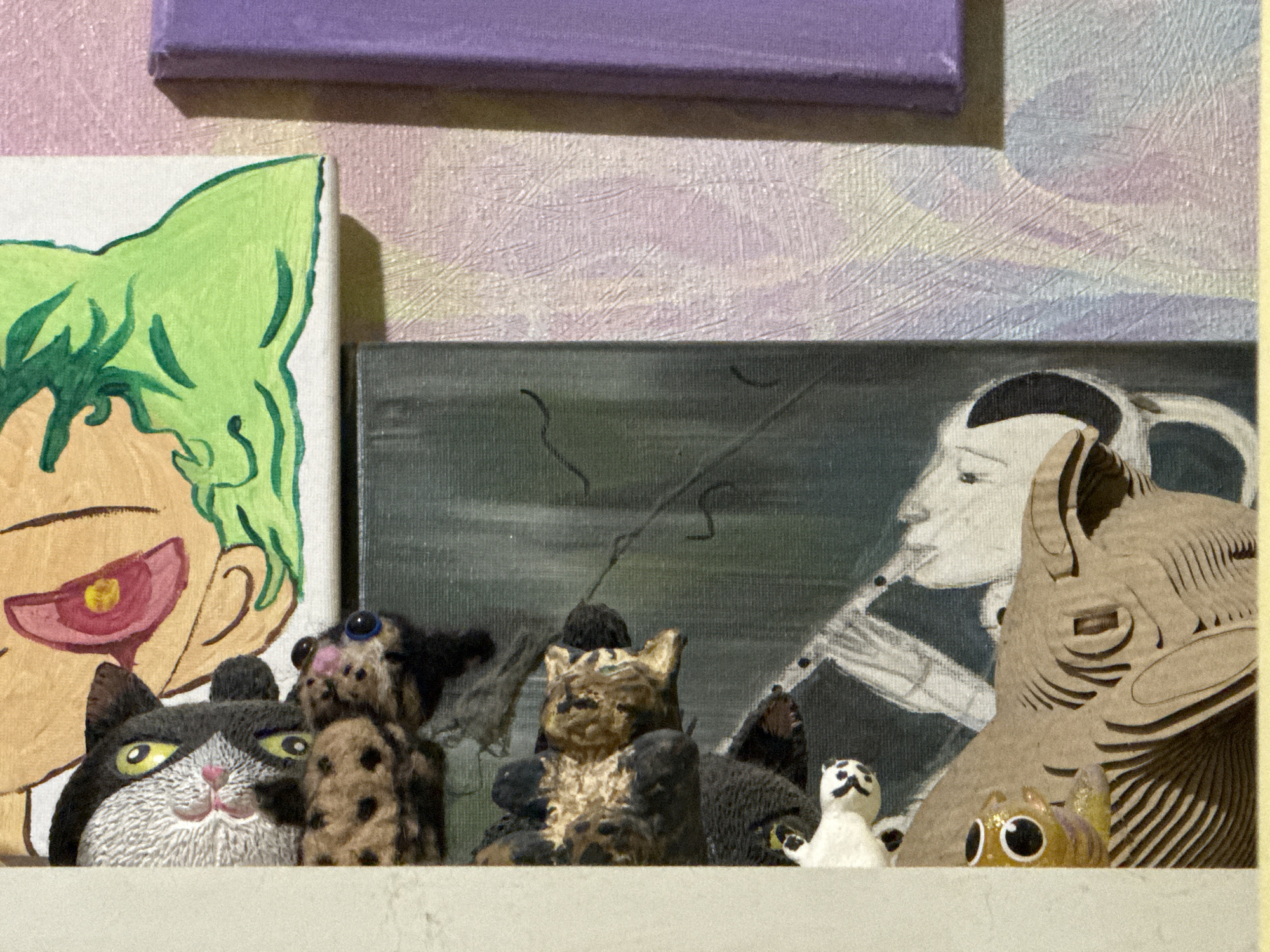 The image displays a collection of small figurines and artwork arranged on a shelf. In the foreground, several cat figurines are visible. From left to right, there is a black and white cat figurine with yellow eyes and a pink nose. To its right, a spotted figurine, possibly a cat, with blue eyes is partially obscured. Next, a tan and black striped cat figurine is positioned centrally, with dark brown figurines on either side. Further to the right, a small white figurine with black markings and a dark figure are present. A small, translucent orange figurine with large black and white eyes is in front of a larger, light brown, layered cardboard cutout of a stylized animal head with visible layers. In the background, two canvases are visible. The canvas on the left features a drawing of a person's face with green hair and a red and yellow eye. The canvas on the right appears to be a painting with abstract shapes and figures, possibly depicting a profile of a person with dark hair. Above these canvases, a purple rectangular object, possibly a book or a mat, is positioned horizontally. The background wall has a textured surface with a pattern of faint lines in pastel colors, predominantly pink and white. The shelf itself is a plain, light-colored surface. There is no discernible information indicating a specific location within ЖК «Борисоглебское», Russia.