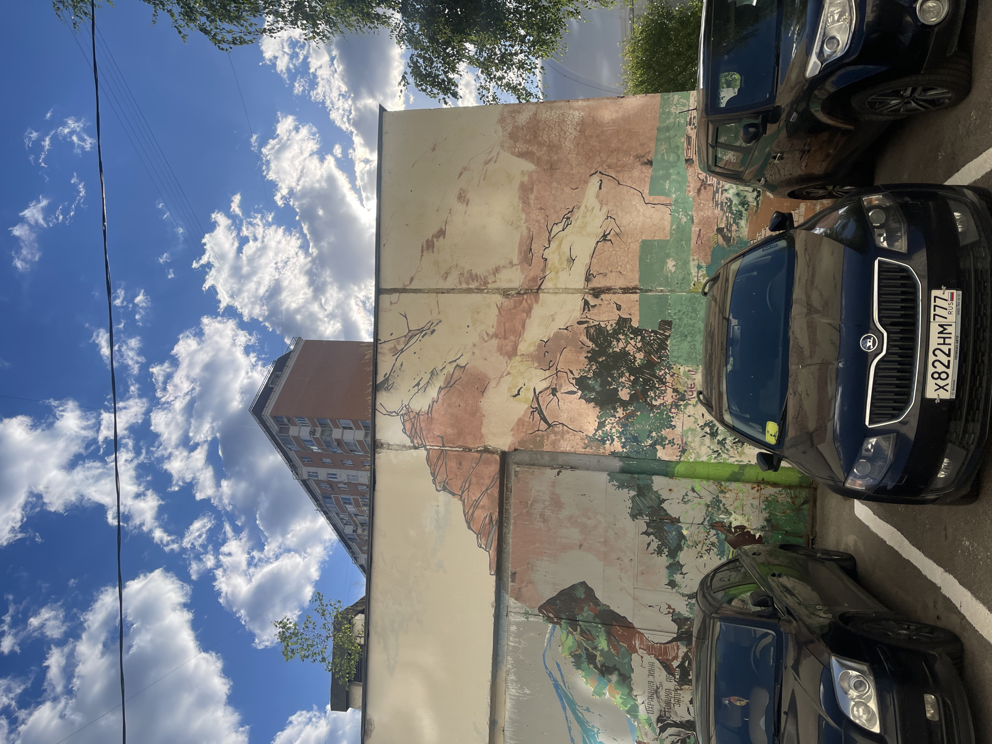 This is a vertical, medium-shot, outdoor photograph taken on a sunny day in Moscow, Russia. The scene features a street art mural painted on the side of a building, with two dark-colored cars parked in front of it.
The mural is abstract and appears to be a landscape or nature-themed painting. It uses earthy tones of beige, brown, and green, with black lines suggesting branches or natural formations. The mural is divided into panels by vertical lines, and some parts are faded or worn. A tall, multi-story brick building is visible in the background on the left side of the frame, against a bright blue sky filled with fluffy white clouds. The sky takes up a significant portion of the left side of the image.
The two cars are parked close together, obscuring parts of the mural. The car in the foreground is a dark sedan, while the car behind it is a dark hatchback or SUV. The license plate on the rear of the front car is visible and reads "822HM 777 RUS". A sticker with Cyrillic text is visible on the front windshield of the sedan.
The time of day appears to be daytime, likely late morning or afternoon, given the bright sunlight. The weather is clear and sunny, as indicated by the blue sky and prominent white clouds. The setting is an urban environment, likely a street or parking area adjacent to a building with a mural. There are no people visible in the image. The overall impression is one of a typical urban scene on a pleasant day.