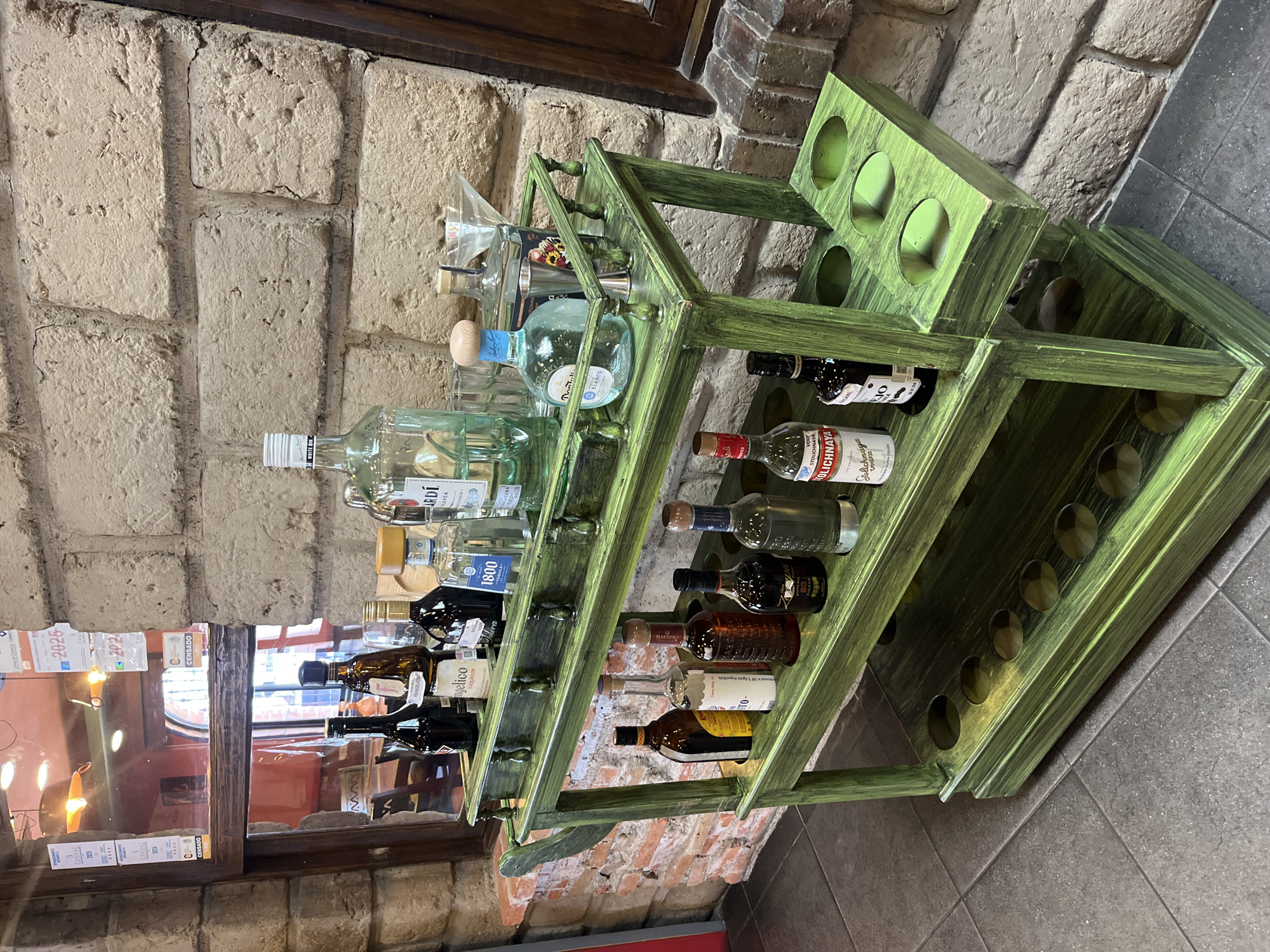 The image captures a green wooden liquor rack stocked with various alcoholic beverages. The rack, appearing aged with distressed paint, is set against a backdrop of a textured brick wall. The location is suggested to be in Puebla, Mexico, based on details observed in the surrounding environment.
The liquor bottles vary in shape and size, including brands like 1800, Maestro Dobel Blanco, and Stolichnaya. Above the rack are some glasses and barware. In the background, there is a window with small posters. The flooring is tiled, adding to the setting's overall texture and visual interest. The image is slightly tilted, creating a dynamic angle in the composition.
